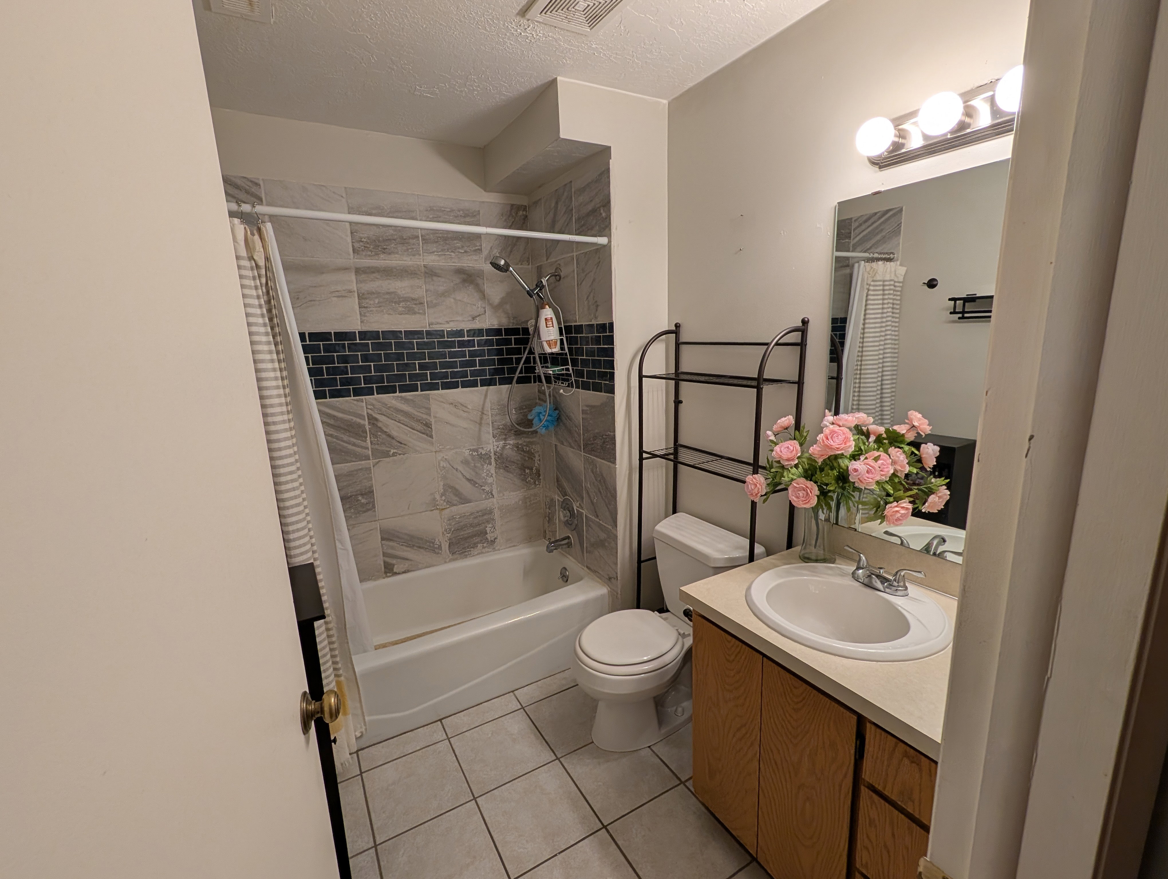 Bathroom featuring a white tub, shower with light gray and dark blue mosaic tile surround, wood-finish vanity with an integrated sink, and tile flooring