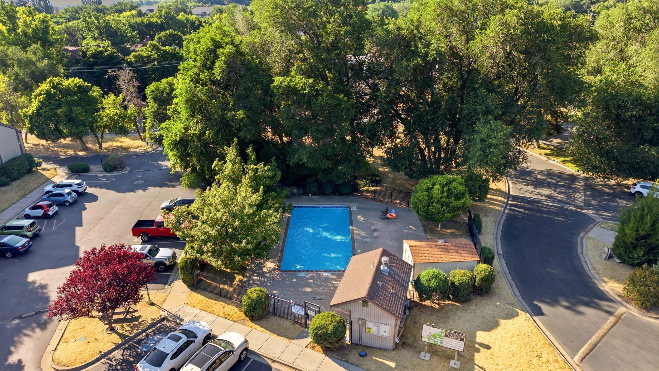 Outdoor swimming pool surrounded by mature trees, featuring a nearby building with a shingled roof, paved parking areas, and surrounding green landscaping
