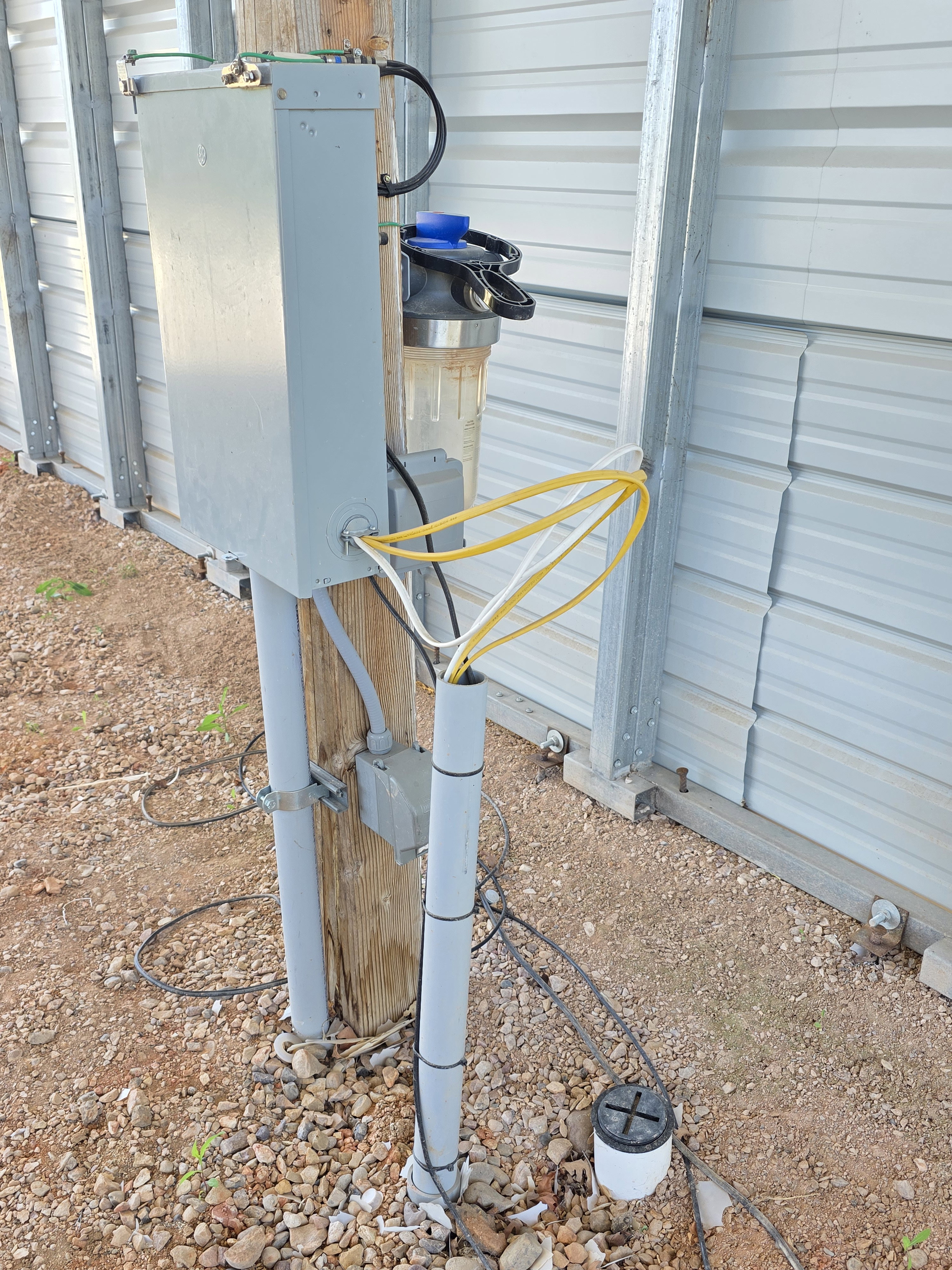 Exterior utility installation featuring a grey electrical panel, clear water filter, and conduit piping on a wooden post