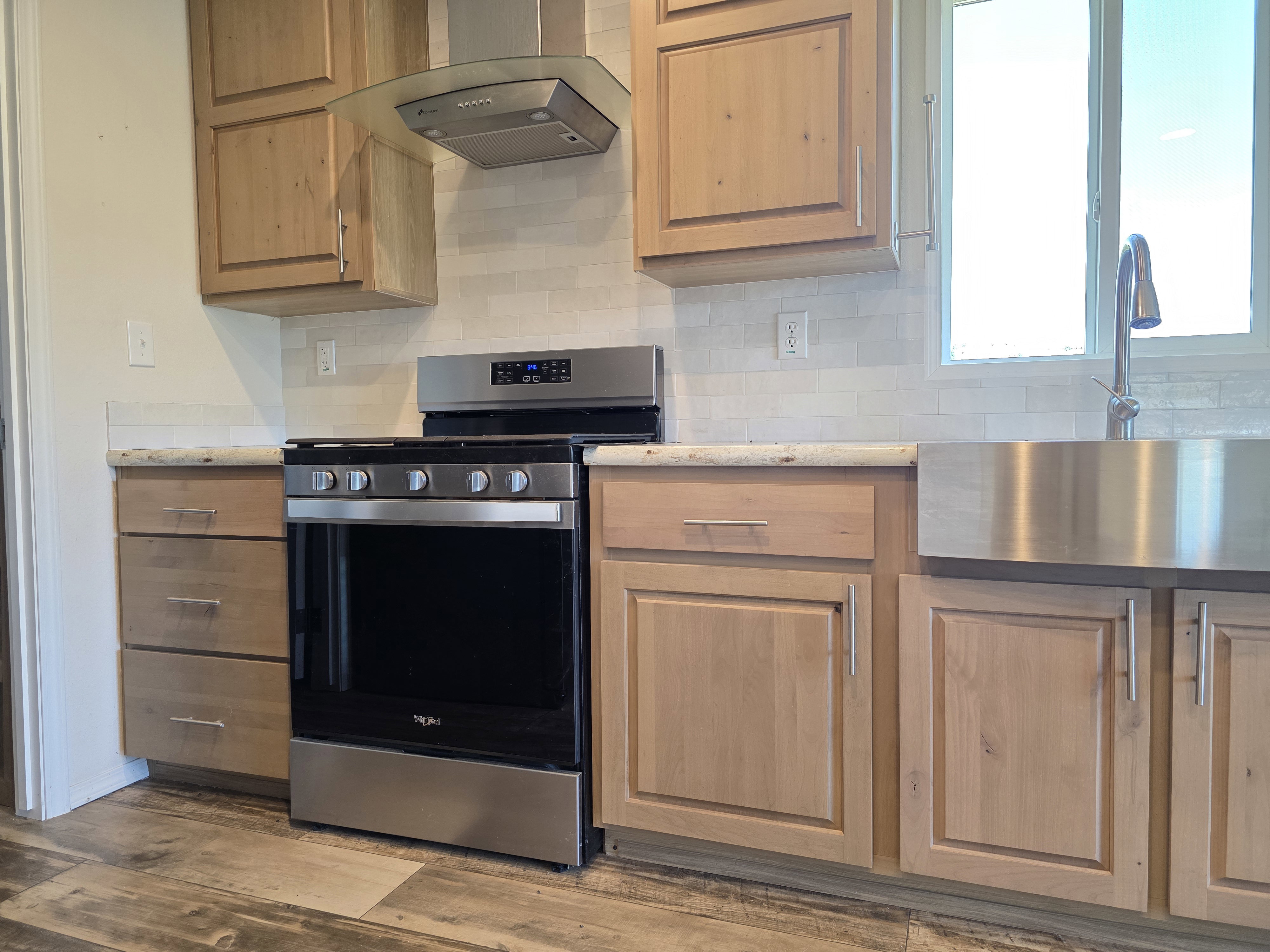 Kitchen featuring natural wood cabinetry, stainless steel appliances, a tile backsplash, light-toned countertops, and a farmhouse-style sink