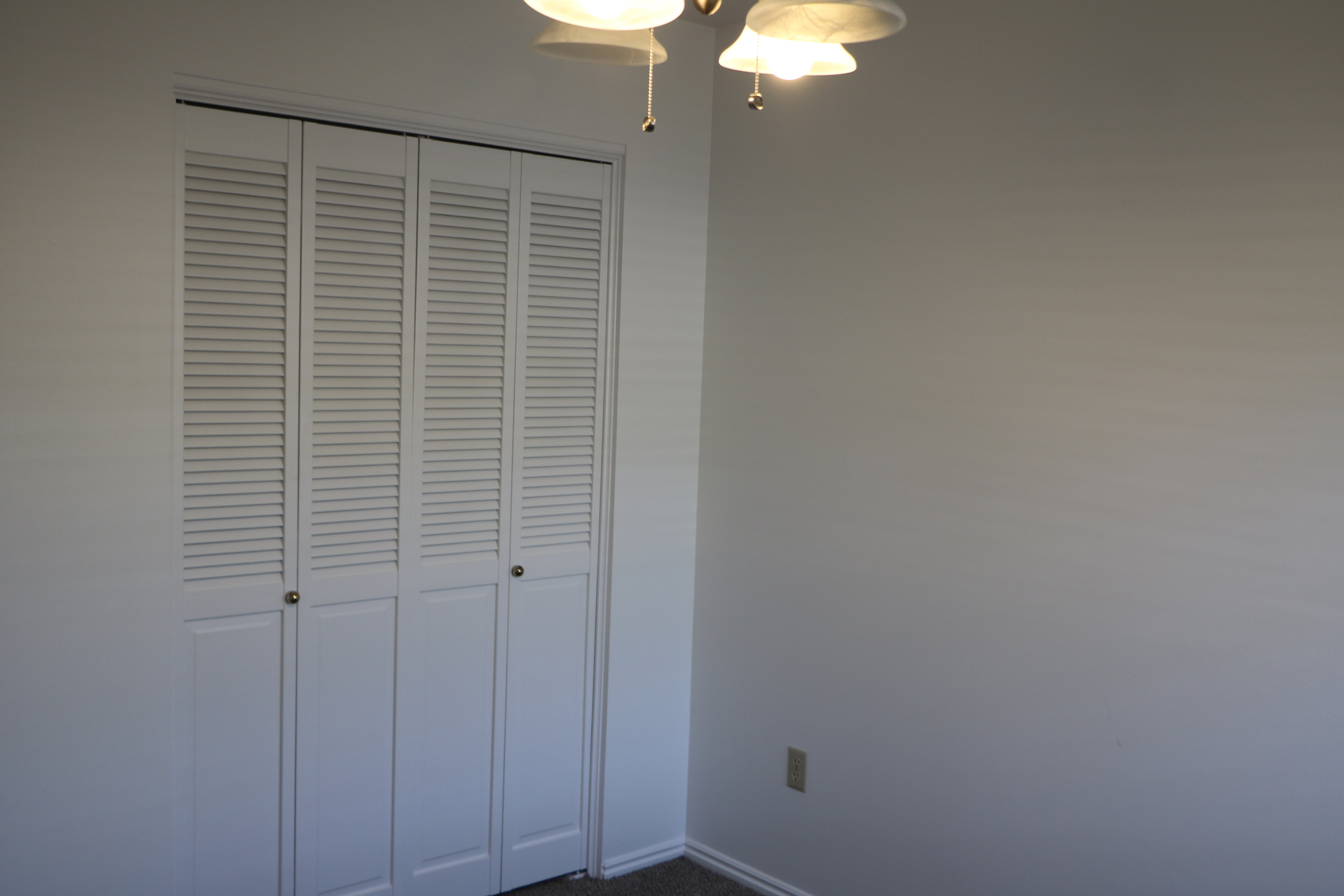 Wall-to-wall louvered bi-fold closet doors with brass-finish hardware, light neutral wall paint, and a ceiling-mounted light fixture with frosted glass shades