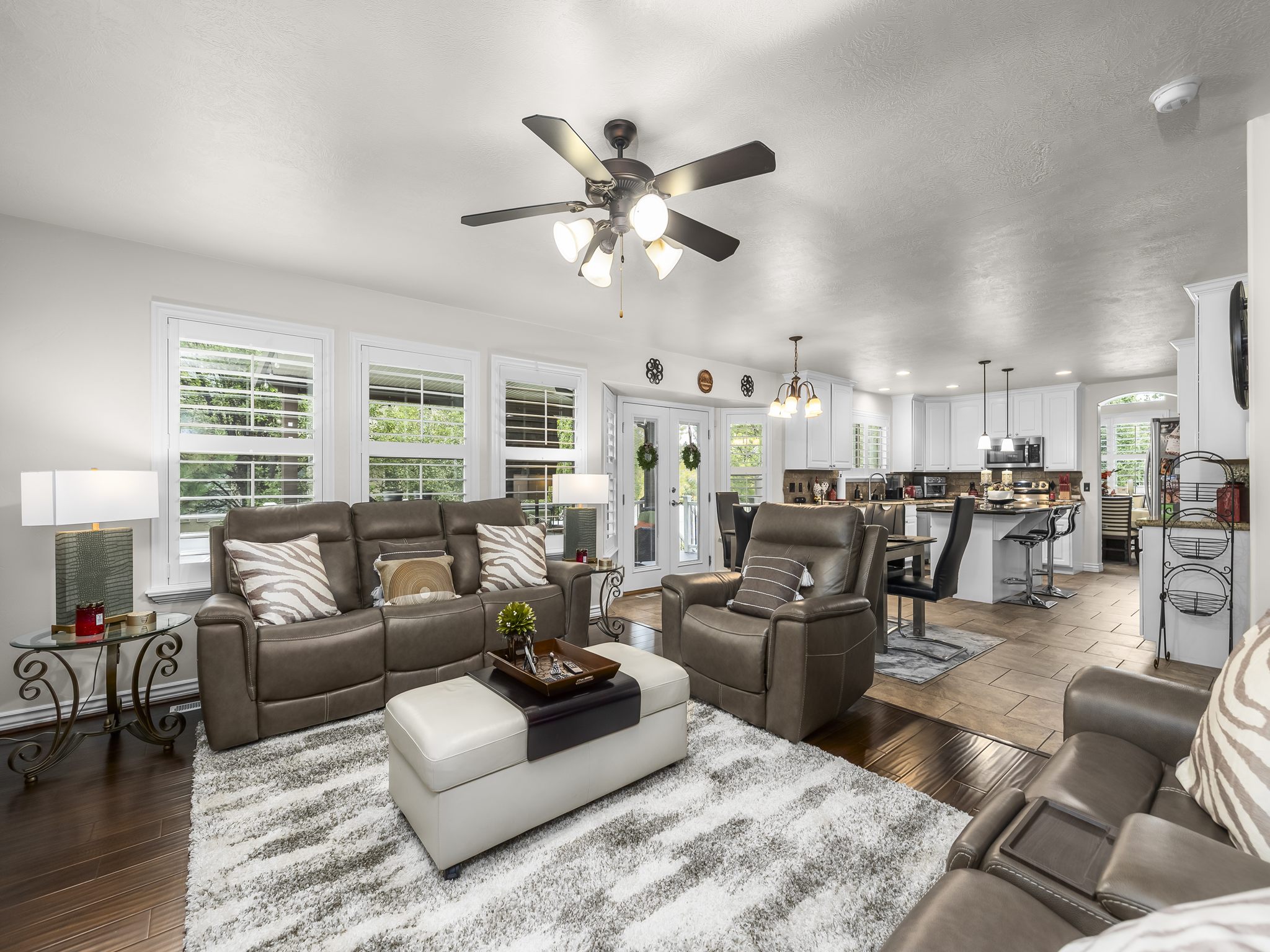 Open-concept living space featuring wood-finish flooring, a ceiling fan with integrated lighting, and multiple windows with plantation shutters
