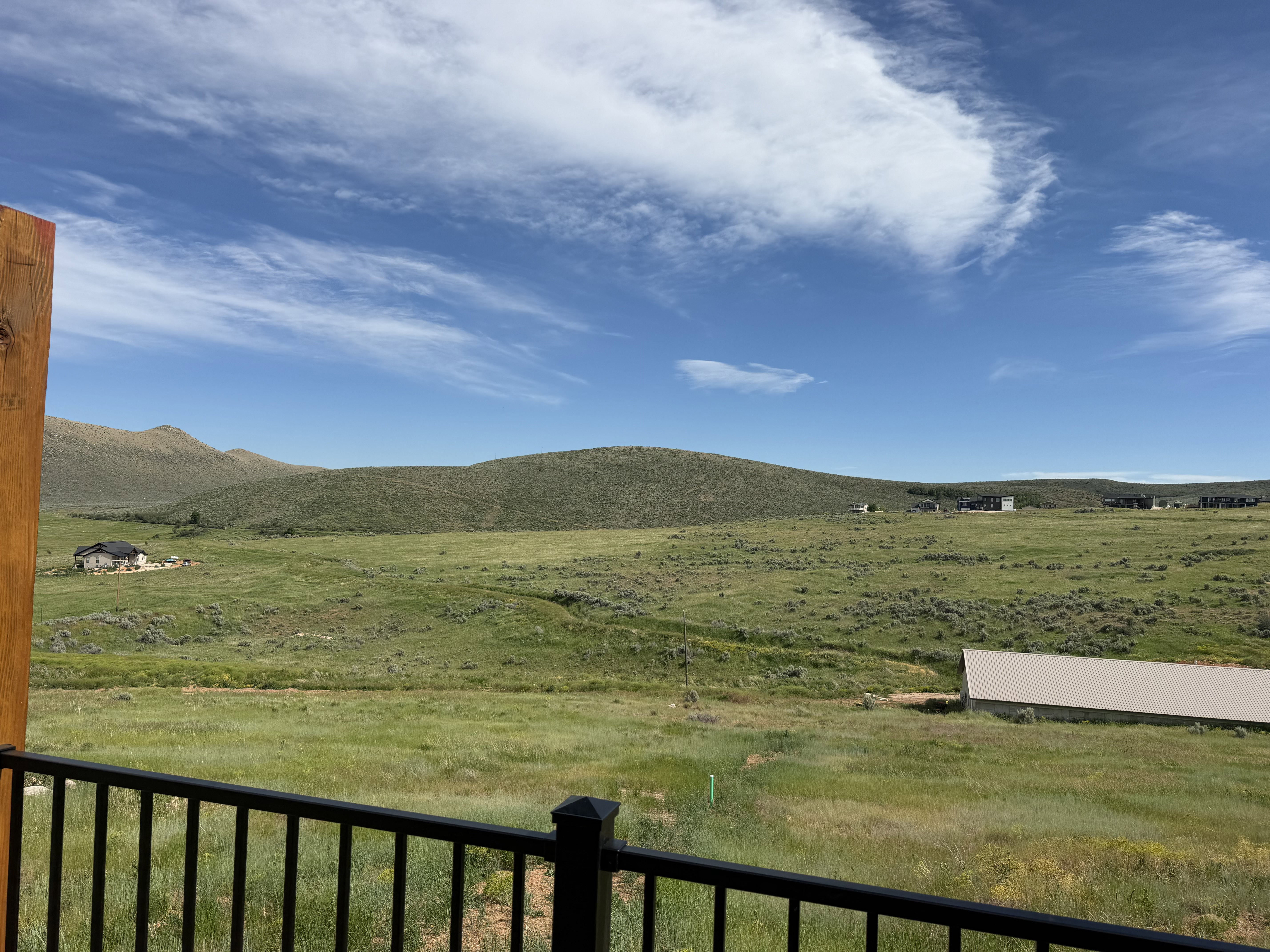 Expansive natural landscape featuring rolling hills and open fields, framed by a dark metal railing and a wood post