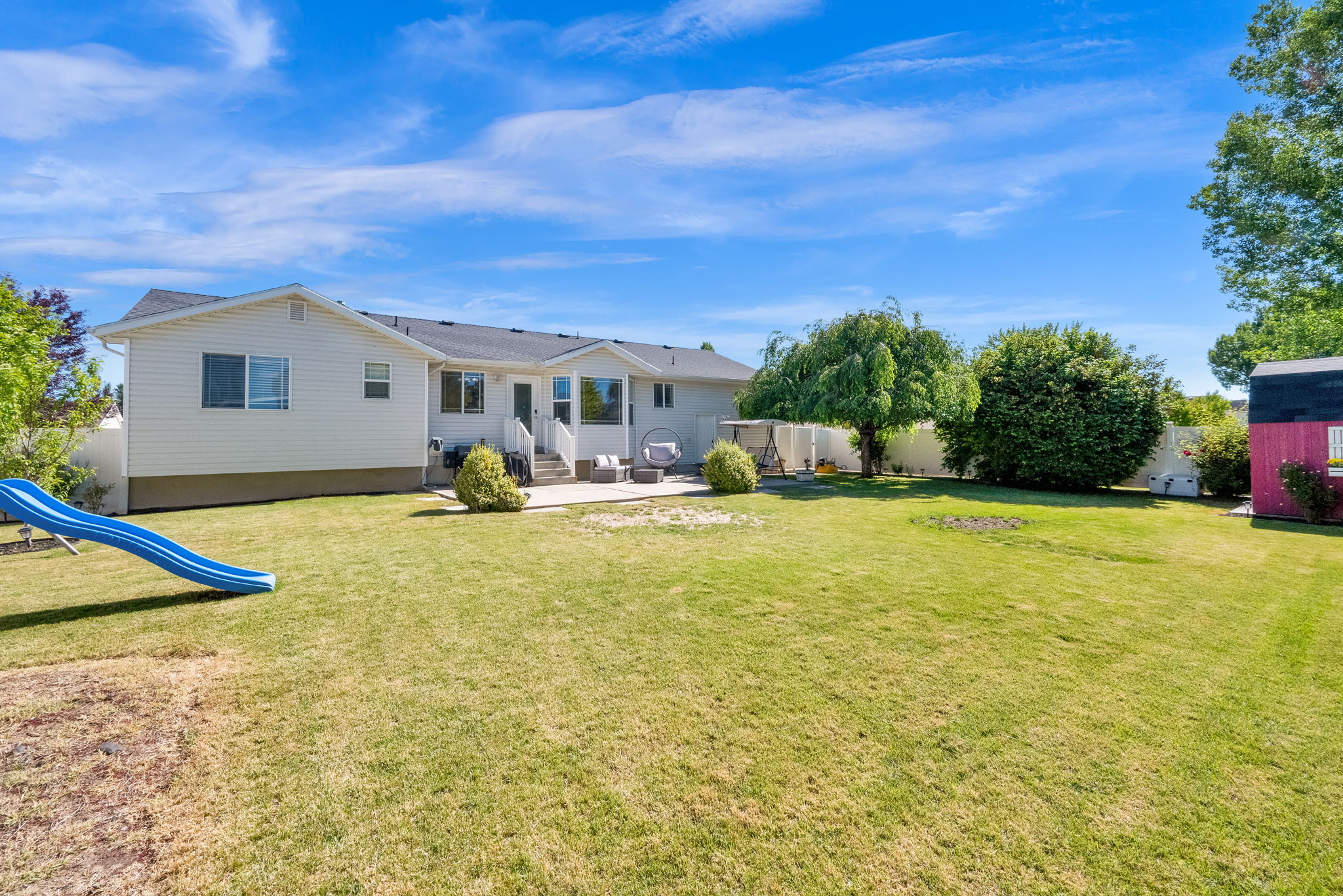 Expansive grassy backyard featuring a concrete patio, mature trees, white vinyl fencing, and a detached storage shed