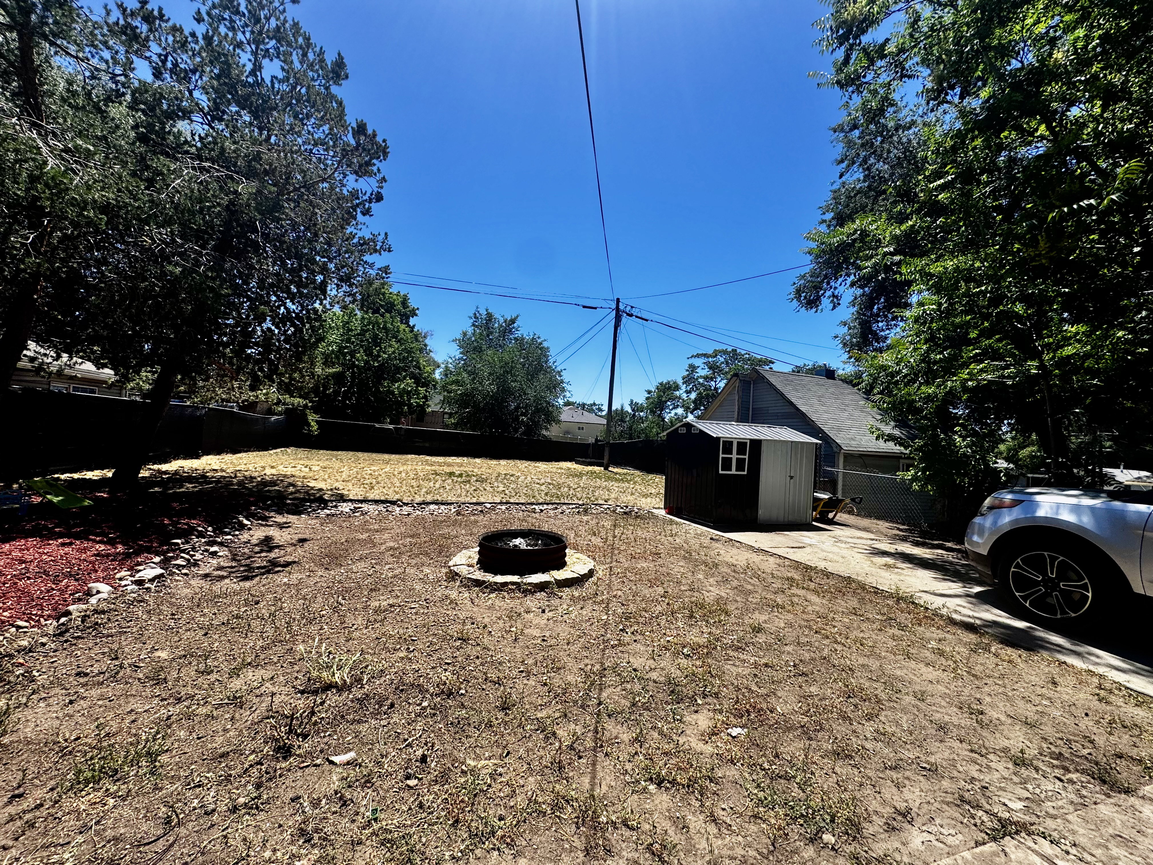 Expansive backyard featuring a built-in fire pit, mature trees, a storage shed, and a paved driveway