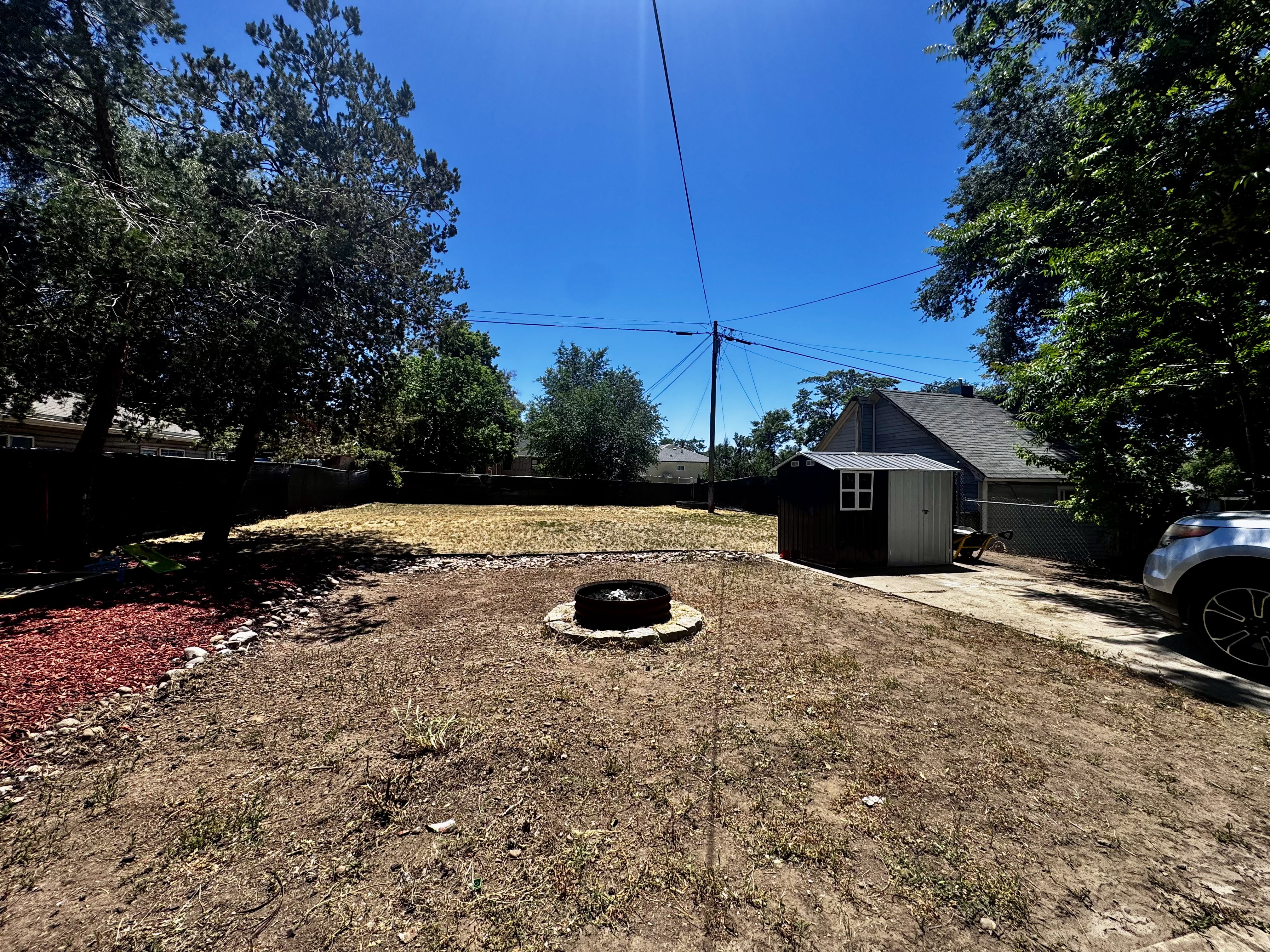 Spacious backyard featuring a stone-ringed fire pit, mature trees, a storage shed, and an expanse of open ground