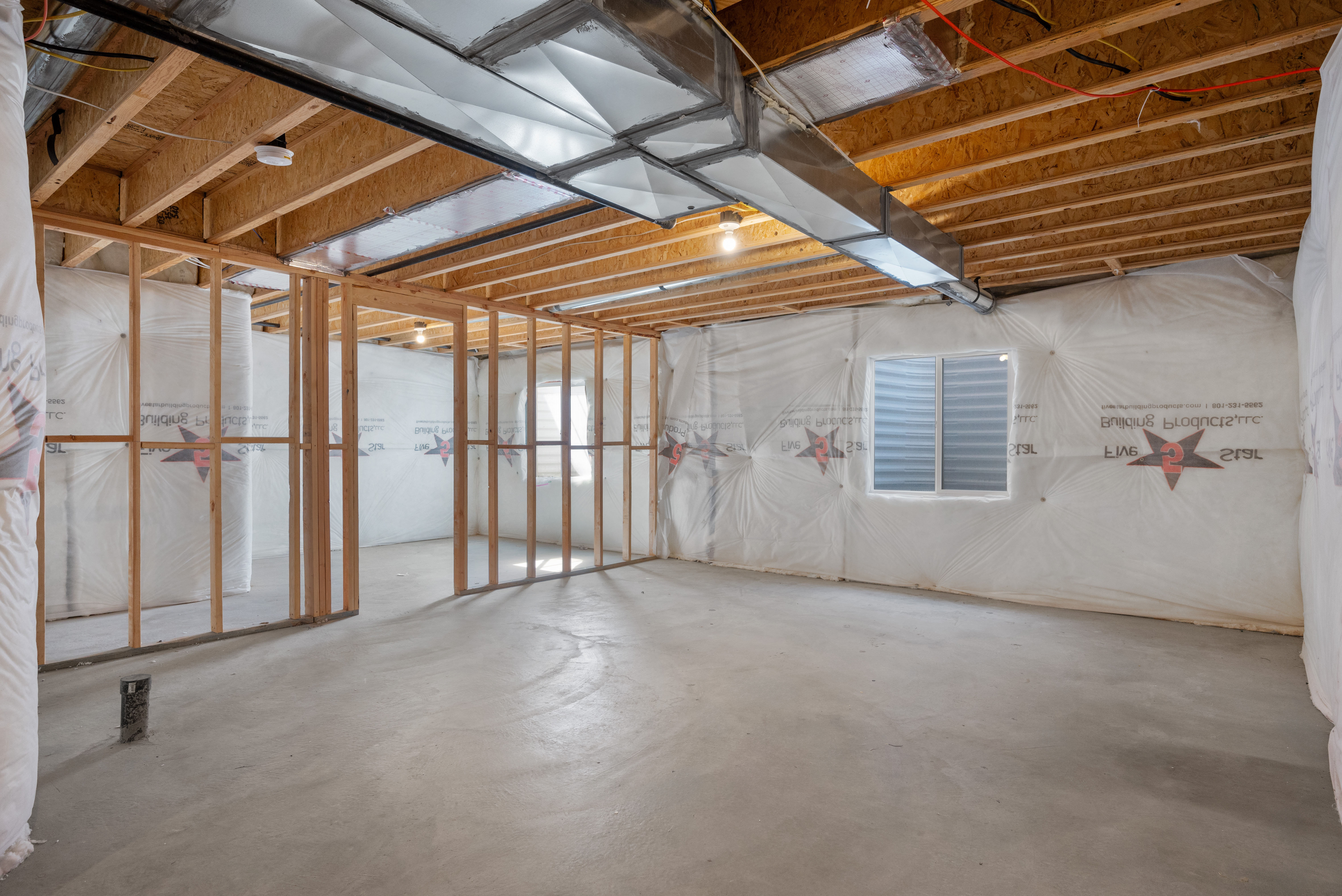 Unfinished basement space featuring exposed wood framing, concrete flooring, a window with an exterior grate, visible ductwork, and insulation-wrapped walls