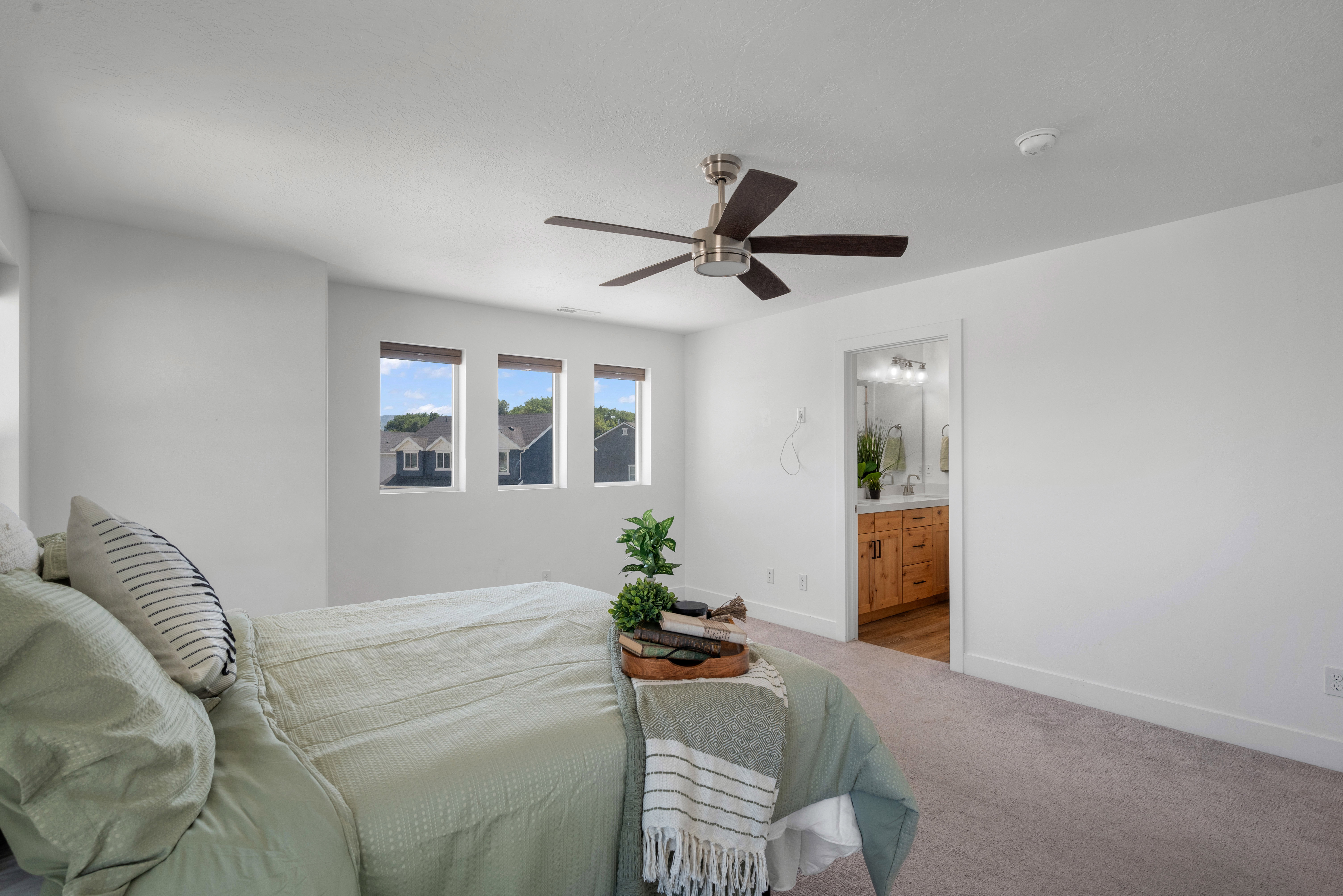 Carpeted room featuring a ceiling fan, three windows, and a doorway revealing a bathroom with a double vanity and recessed lighting