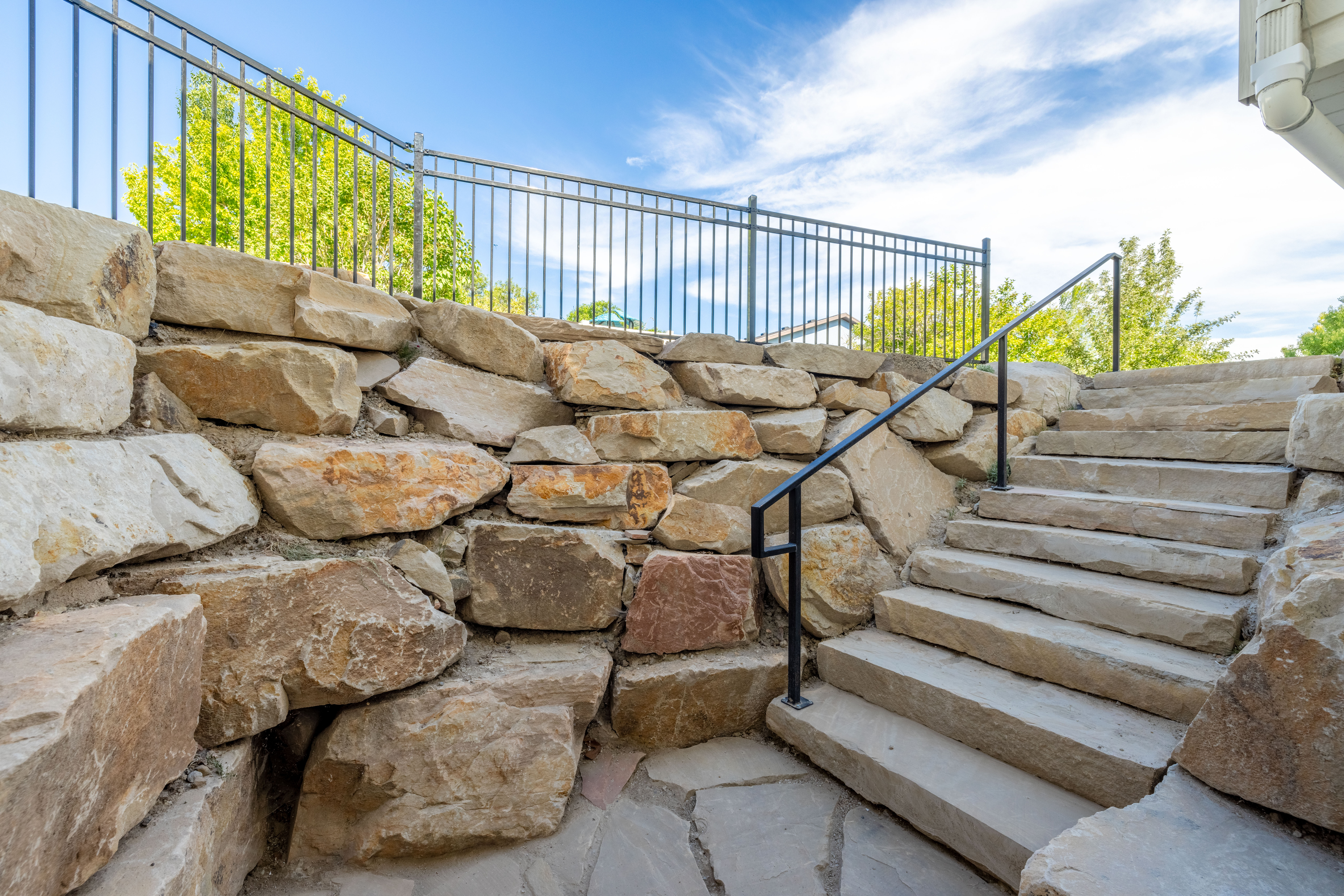 Natural stone steps featuring a black metal handrail and a stone retaining wall