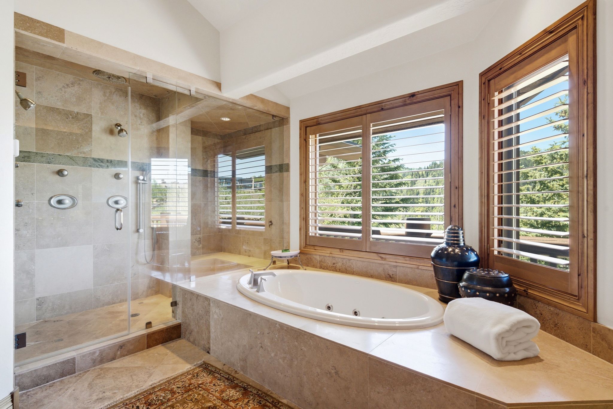 Spacious bathroom featuring a built-in jetted tub, a glass-enclosed shower with body sprays, tile flooring, and wood-framed windows with plantation shutters