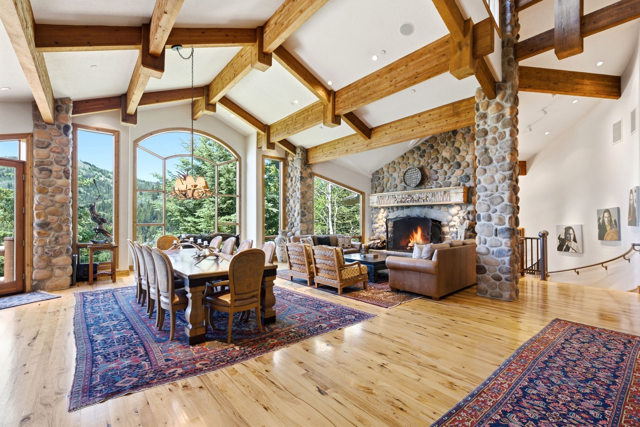Vaulted ceilings with exposed wood beams, a stone fireplace, and wide-plank wood flooring complement the large arched windows