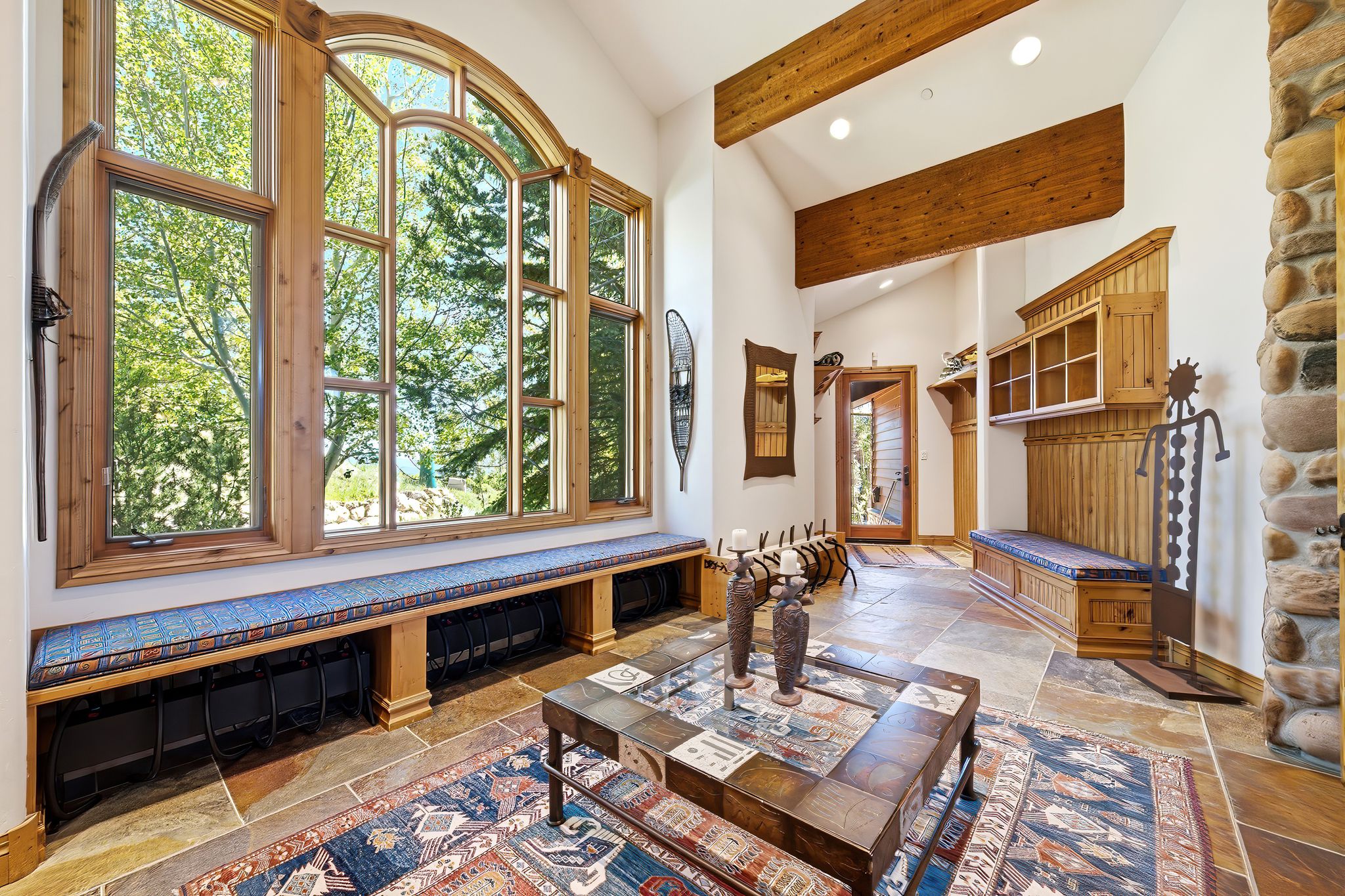Expansive arched window with wood framing, built-in window bench, exposed wood beams, natural stone flooring, and a stone wall accent