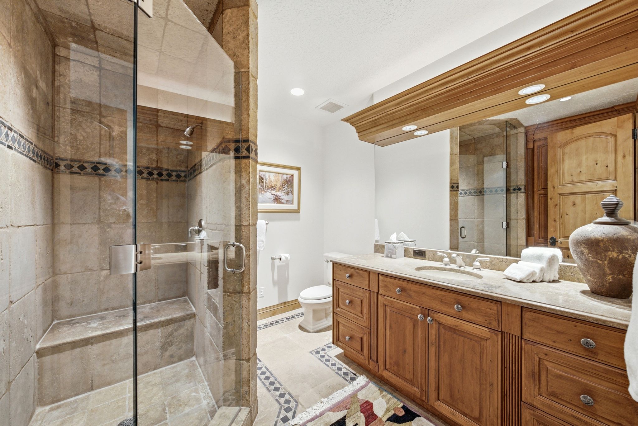 Spacious bathroom featuring a glass-enclosed shower with an integrated bench, extensive wood cabinetry, a large vanity mirror with overhead lighting, a stone countertop, and decorative floor tiling