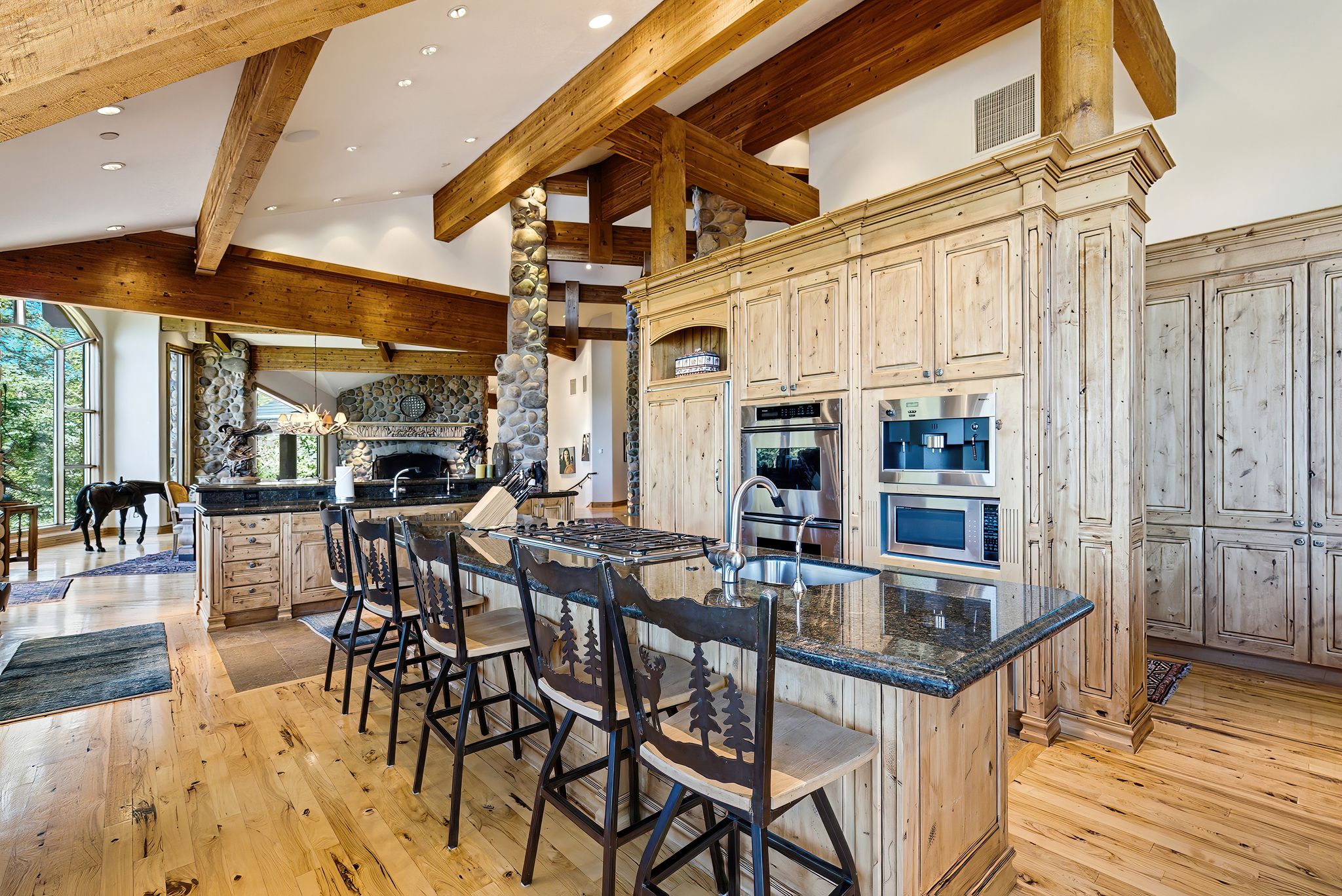 Spacious kitchen featuring an island with a sink and seating, built-in stainless steel appliances, extensive wood-finish cabinetry, exposed wood beams, and hardwood flooring