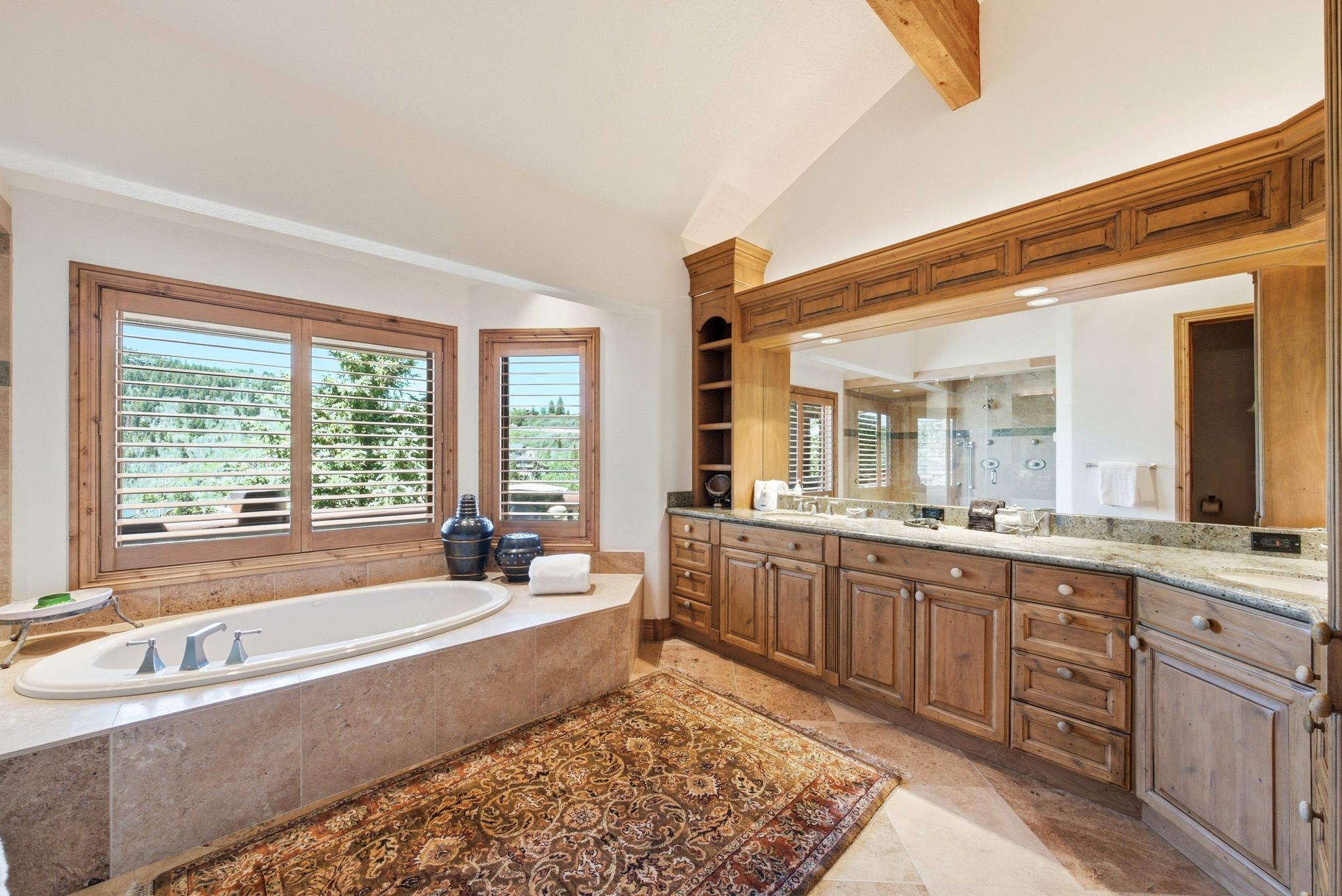 Spacious bathroom featuring a built-in soaking tub with stone surround, custom wood cabinetry, extensive countertop space, wood-finish window frames with plantation shutters, and a beamed ceiling