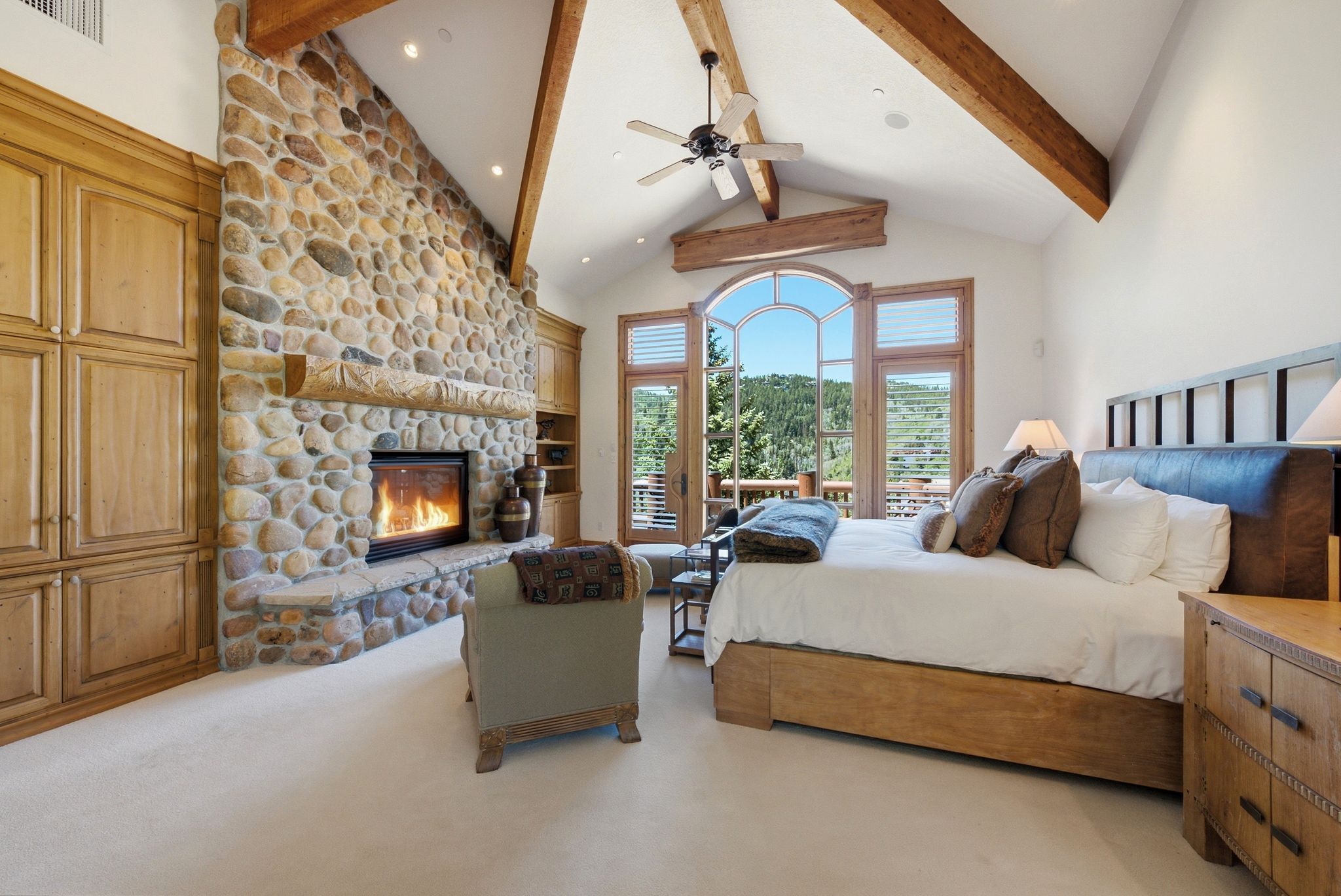 Vaulted ceiling with exposed wood beams, stone fireplace, built-in cabinetry, arched window, and outdoor access