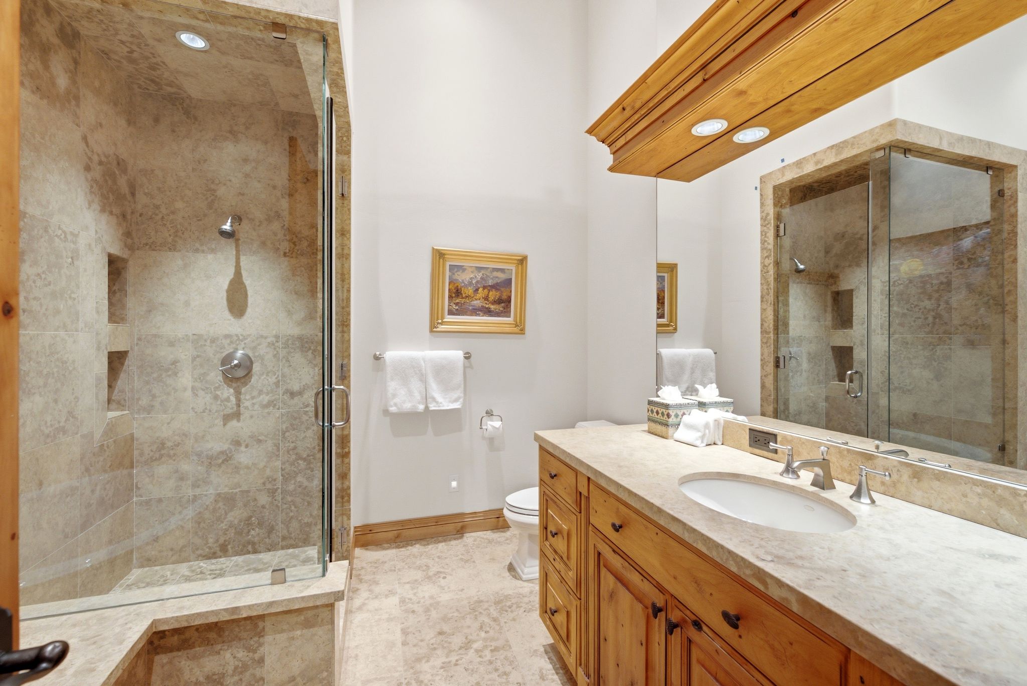 Bathroom featuring a glass-enclosed shower with stone tile surround, a single vanity with wood cabinetry and stone countertop, a large wall-mounted mirror, and stone tile flooring