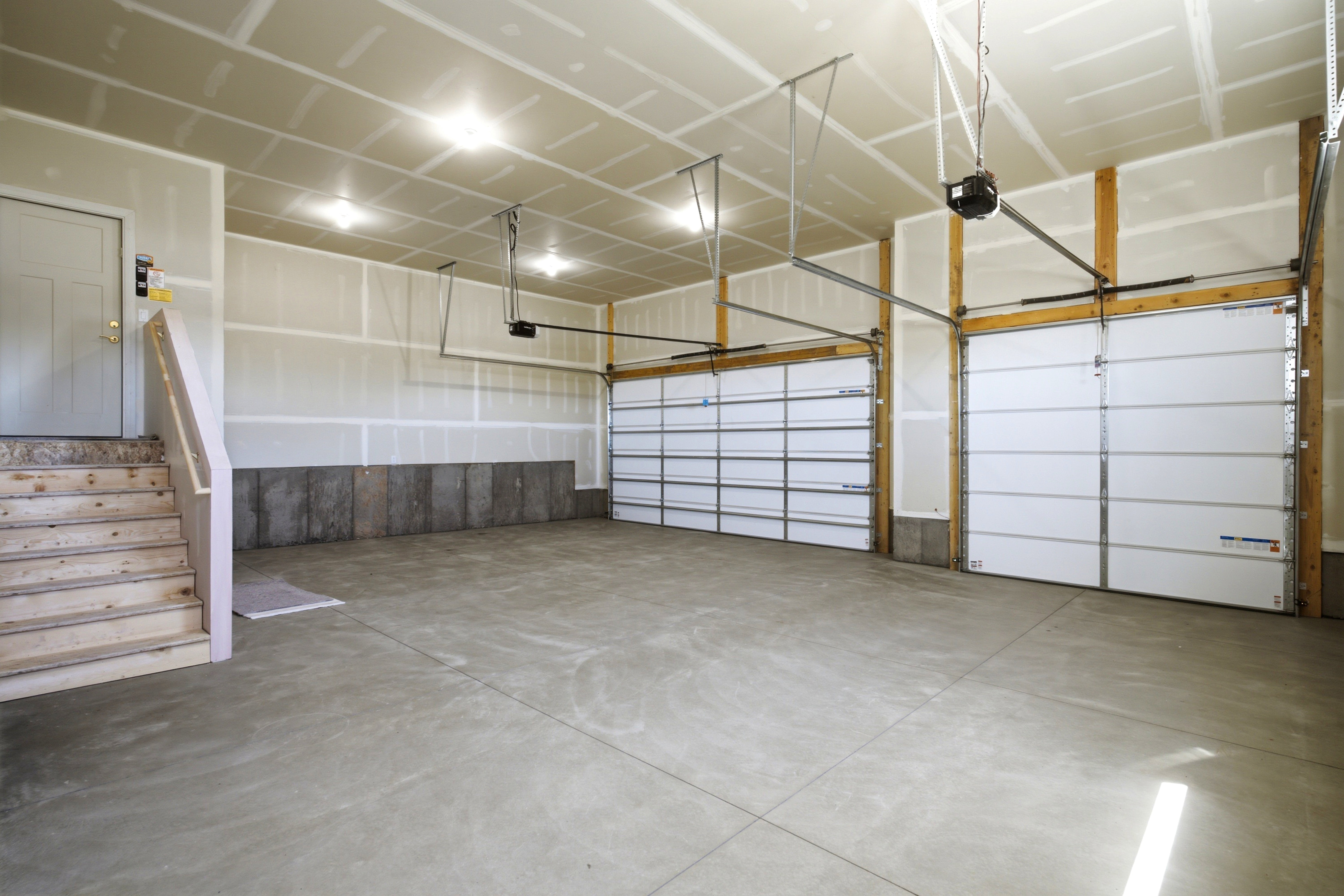 Spacious two-car garage featuring a concrete floor, two white garage doors with openers, and an interior access door with wooden stairs