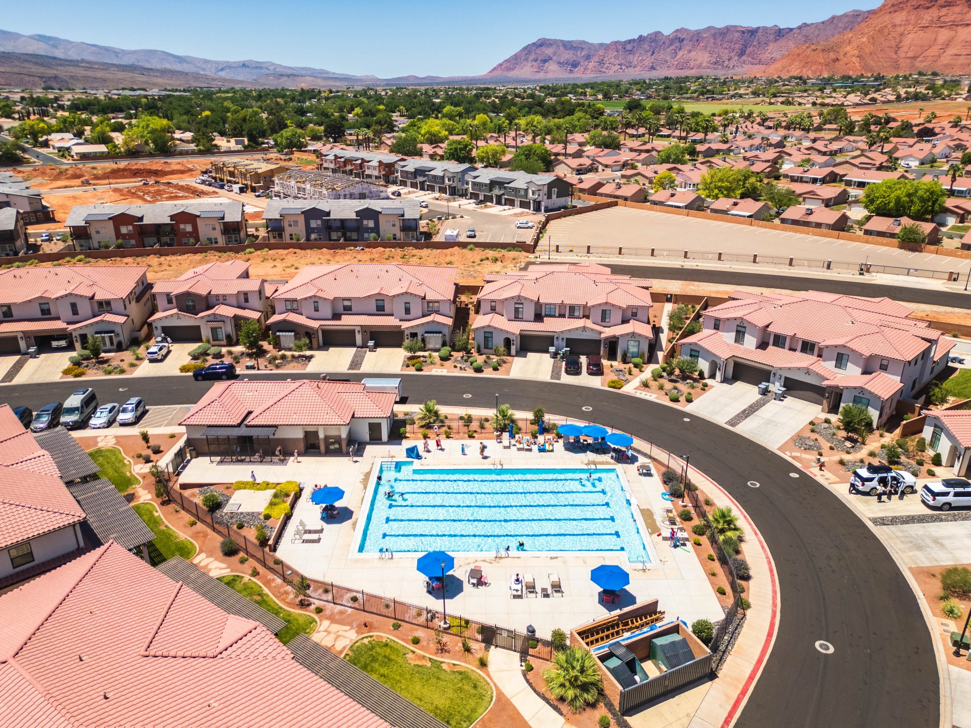 Community swimming pool with surrounding lounge areas and a recreation building, featuring a paved pool deck and blue shade umbrellas