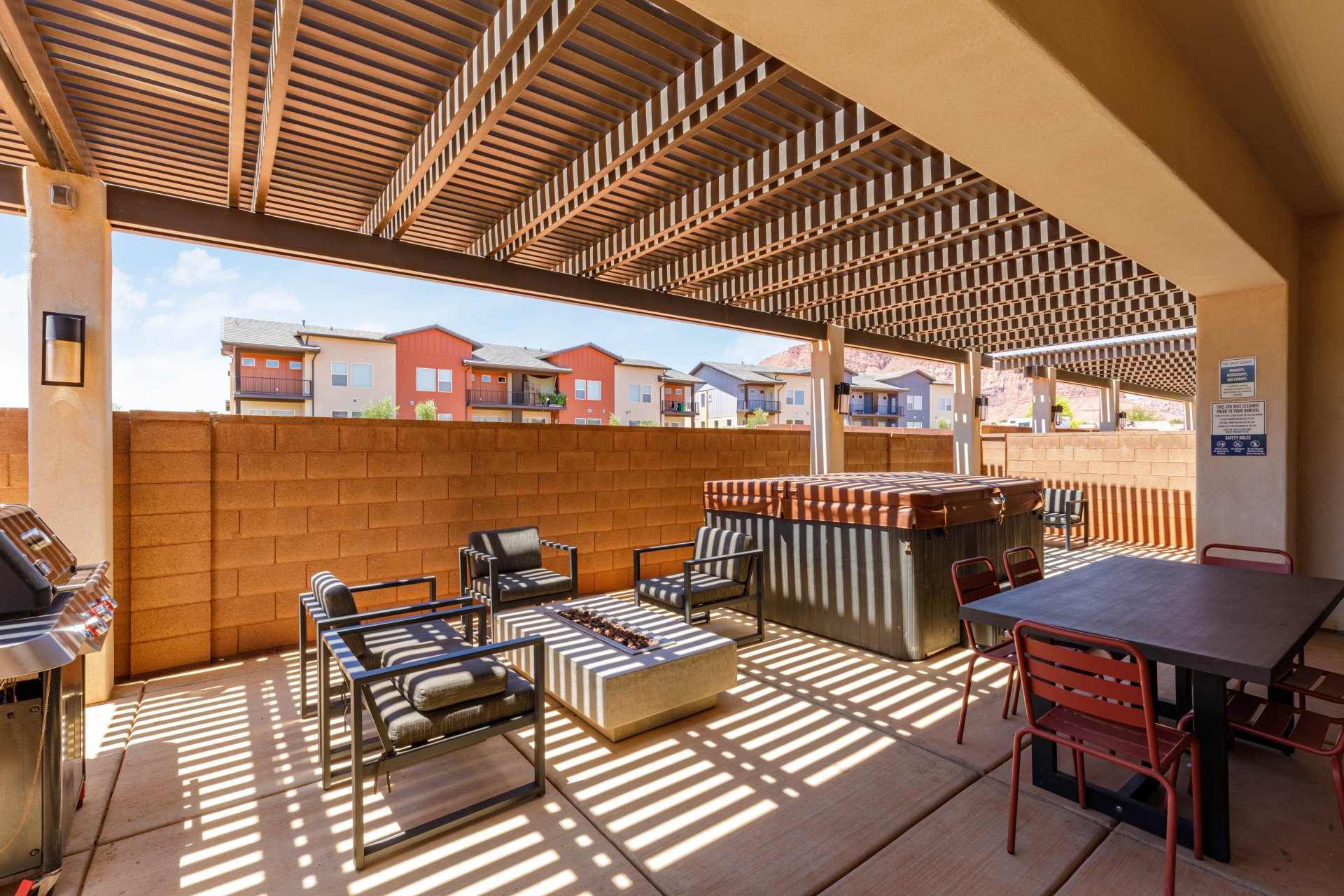 Expansive outdoor patio featuring a slatted pergola, concrete paver flooring, a built-in fire pit, a hot tub, and a durable block wall