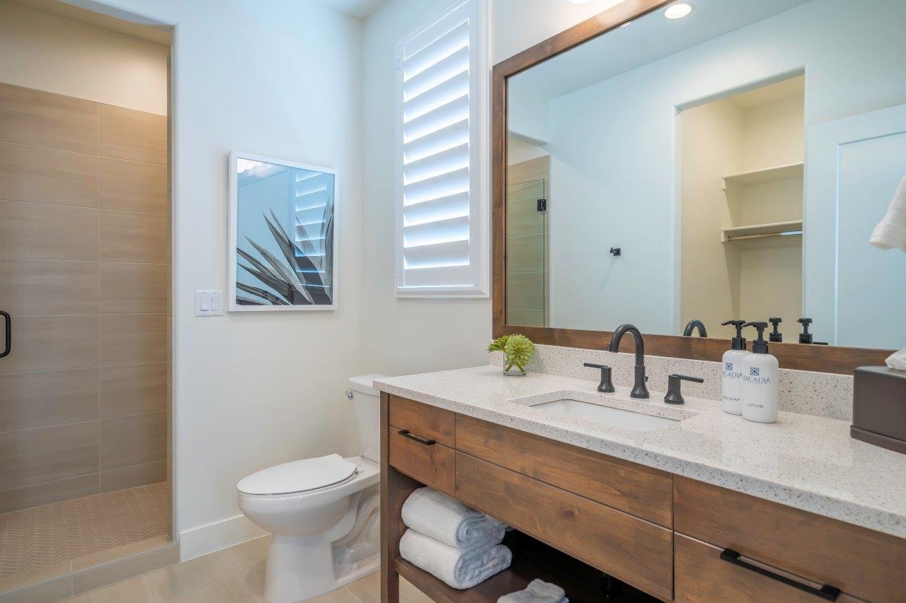 Bathroom featuring a walk-in shower with tile surround, a wood-finish vanity with an integrated sink and light-toned countertop, a large framed mirror, and a window with plantation shutters