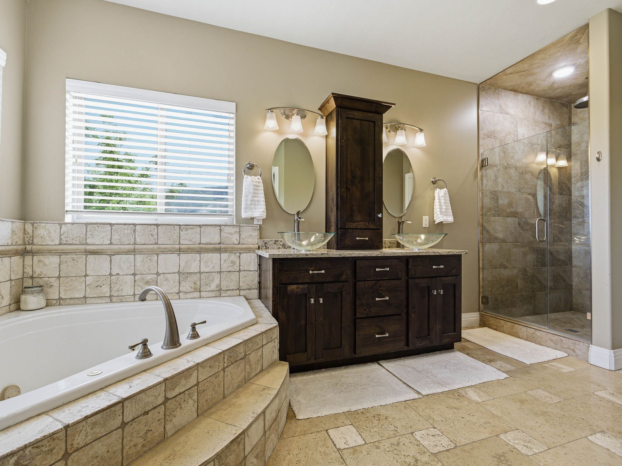 Spacious bathroom featuring a built-in soaking tub, a dual vanity with vessel sinks, a glass-enclosed shower, tile flooring, and a window with horizontal blinds