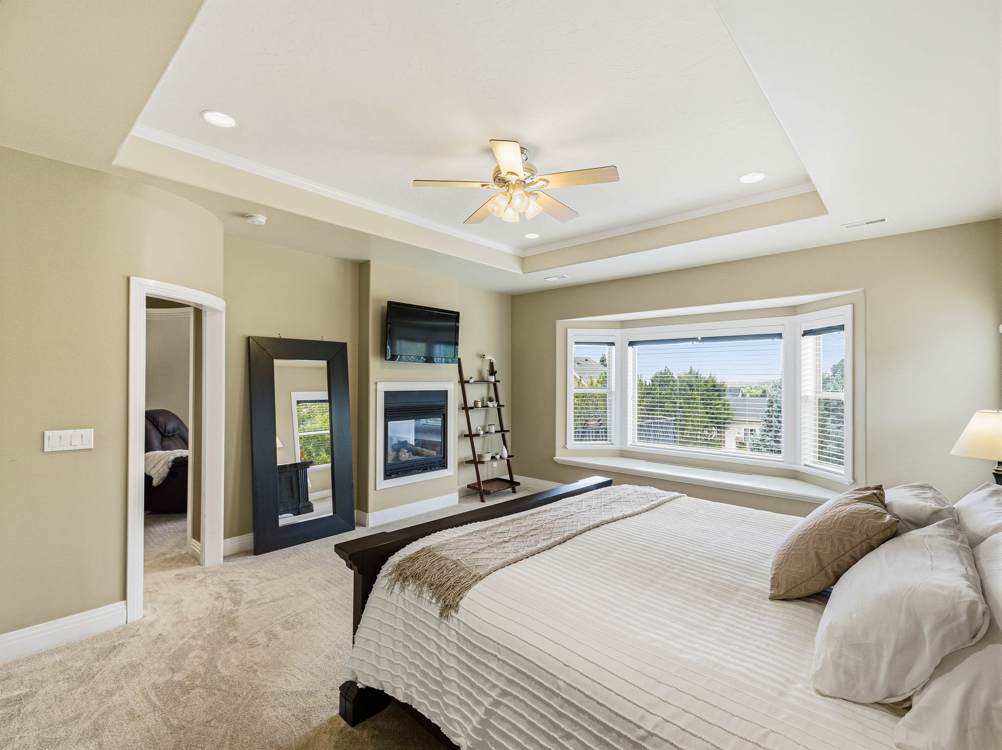 Spacious room featuring a tiered tray ceiling with recessed lighting, a ceiling fan, plush carpeting, a built-in fireplace, and a large bay window with a deep sill