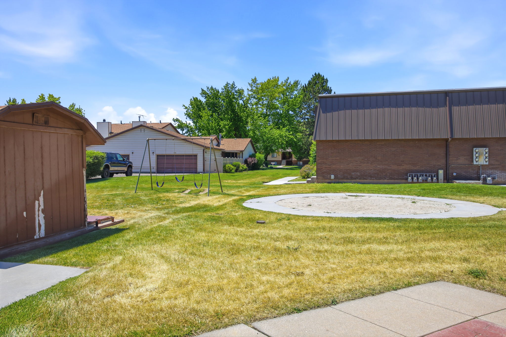 Expansive grassy area featuring a circular sand-covered space, a swing set, and mature trees