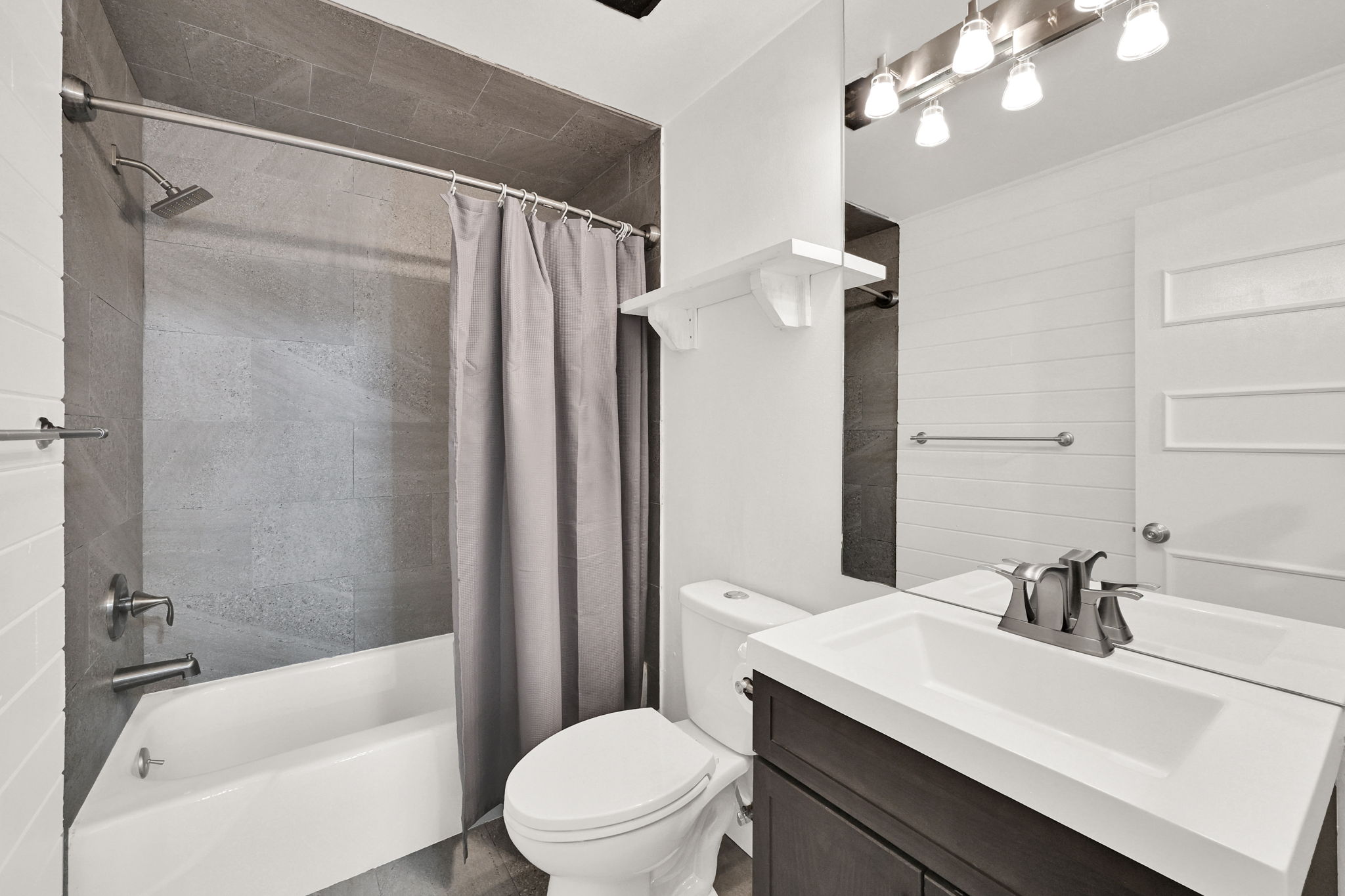 Contemporary bathroom featuring a white tub with gray tiled surround, a dark wood-finish vanity with an integrated white sink, a wall-mounted mirror, and brushed nickel fixtures