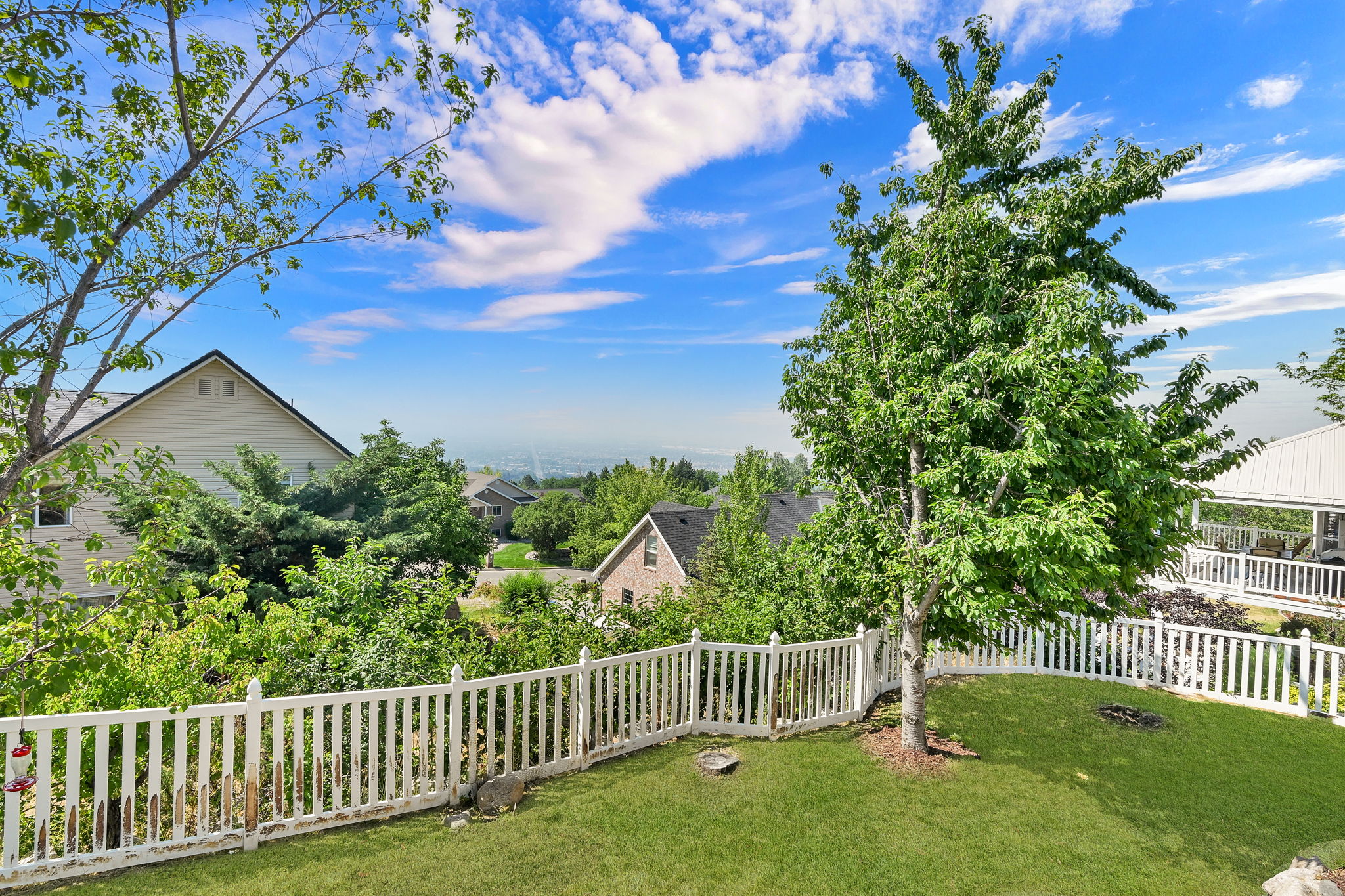 Fenced backyard featuring a manicured lawn, established trees, and a distant cityscape