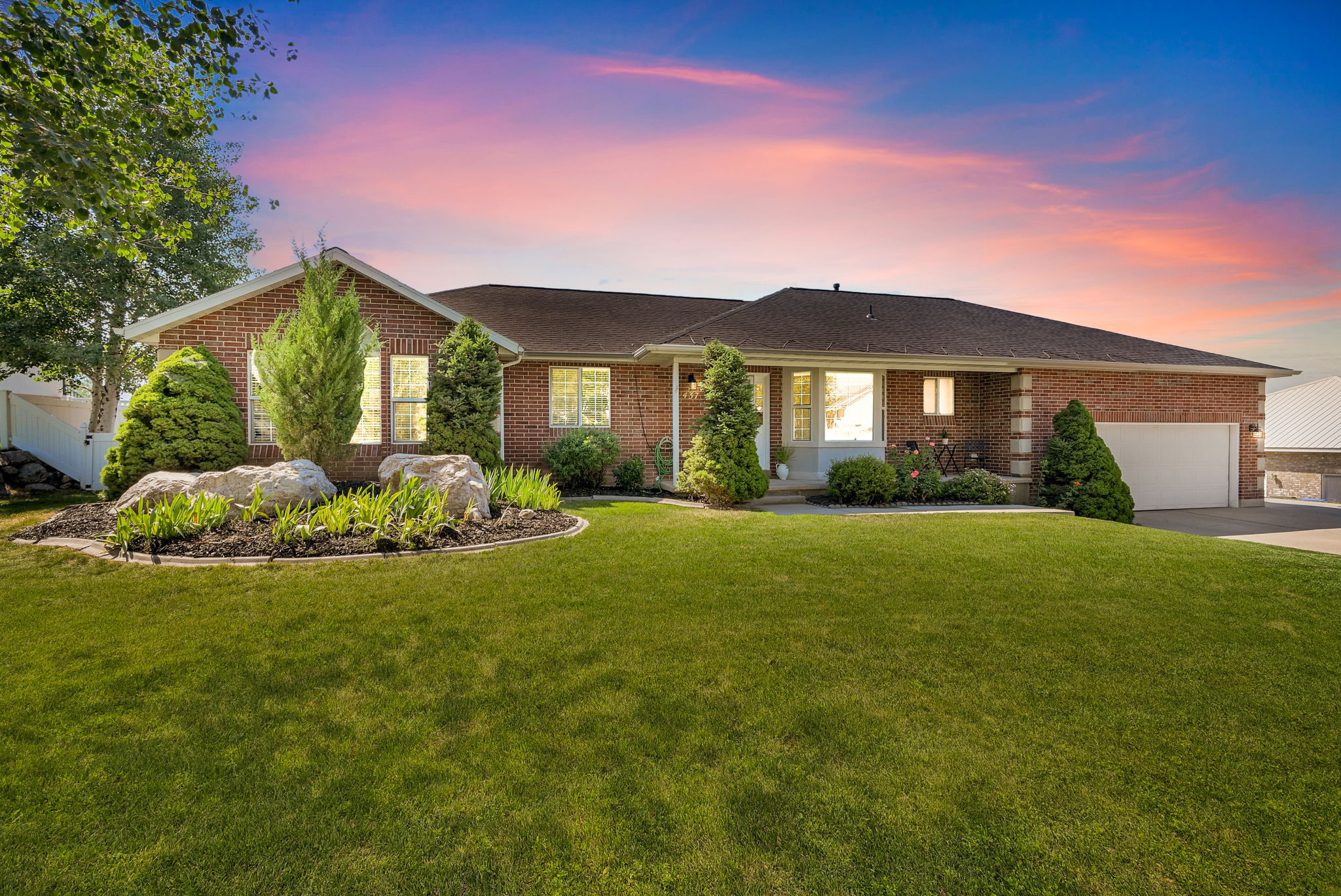 Traditional brick exterior featuring a manicured front lawn, established landscaping with large decorative rocks, a two-car garage with an automatic door, and multiple large windows
