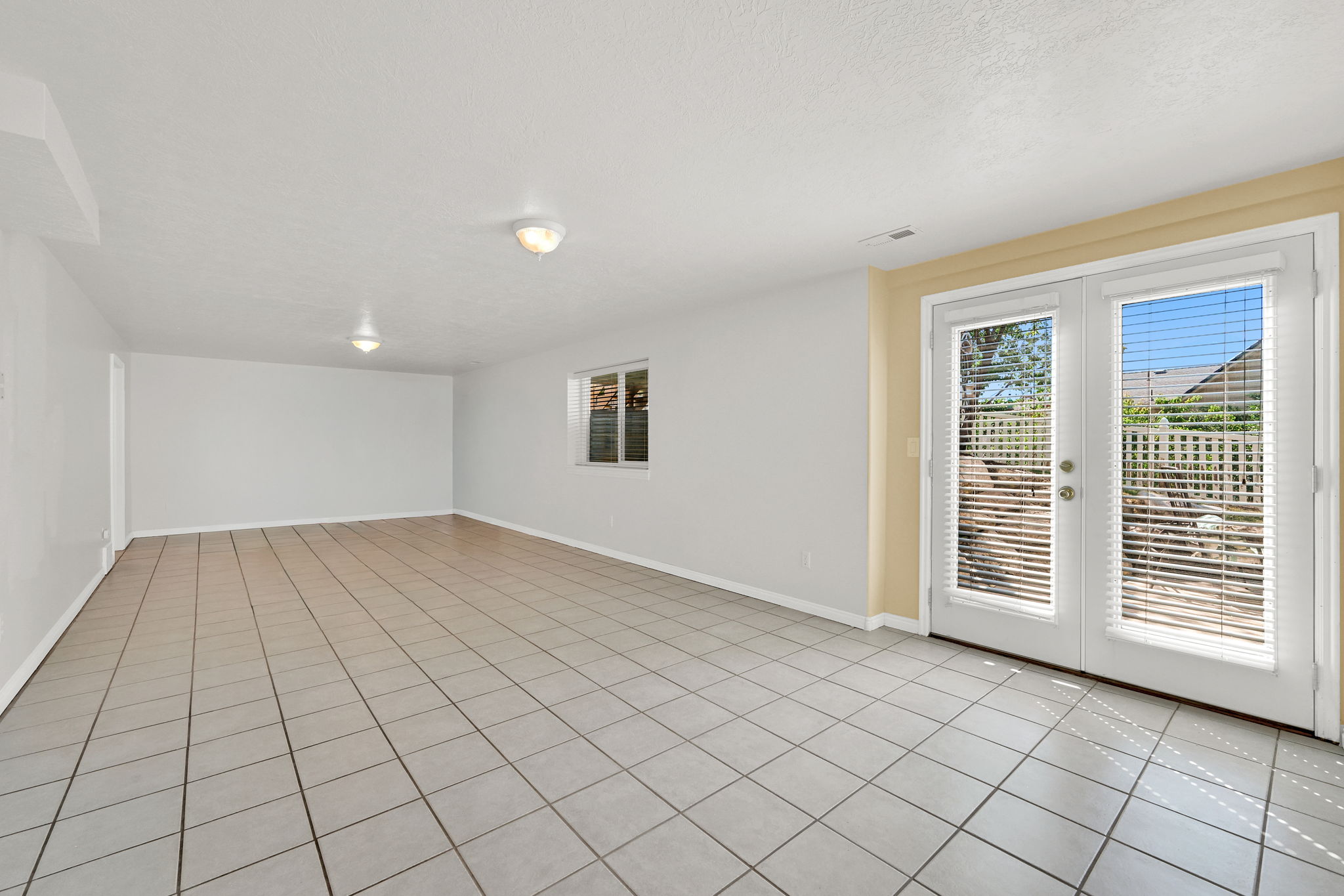 Spacious room featuring light-toned tile flooring, white walls, and a pair of white French doors with integrated blinds