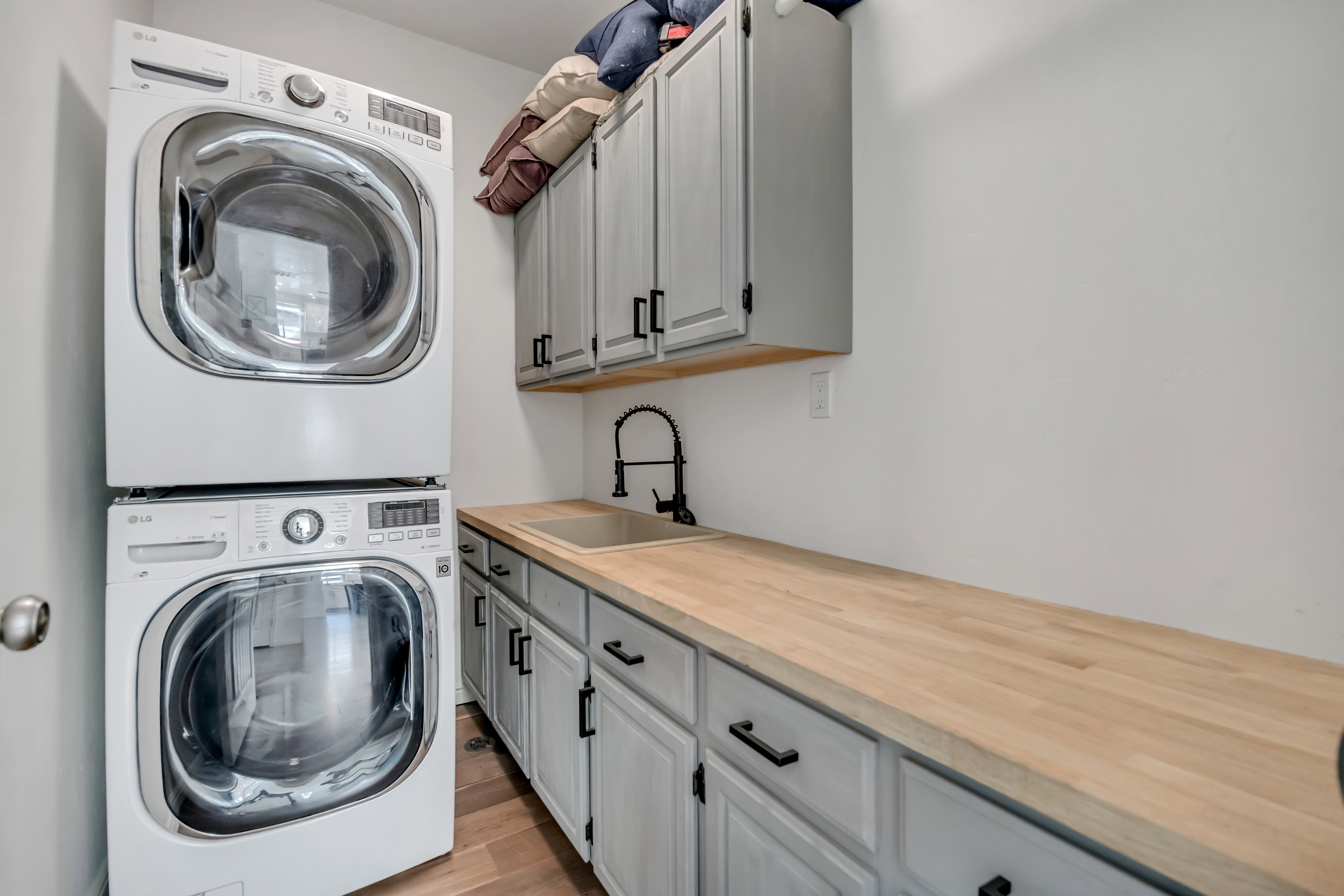 Laundry area featuring stacked front-loading washer and dryer units, light wood-finish countertops, a utility sink with a black gooseneck faucet, and gray cabinetry with black hardware