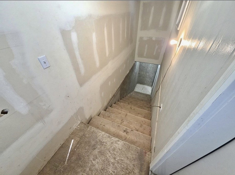 Unfinished lower-level staircase featuring exposed wood treads and risers, drywall-finished walls, and an overhead light fixture