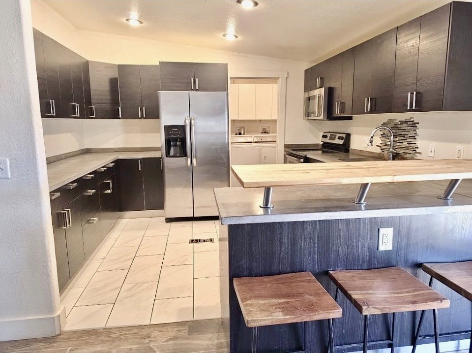 Contemporary kitchen featuring dark wood-finish cabinetry, stainless steel appliances, light-toned countertops, a textured tile backsplash, and a breakfast bar with a wood surface