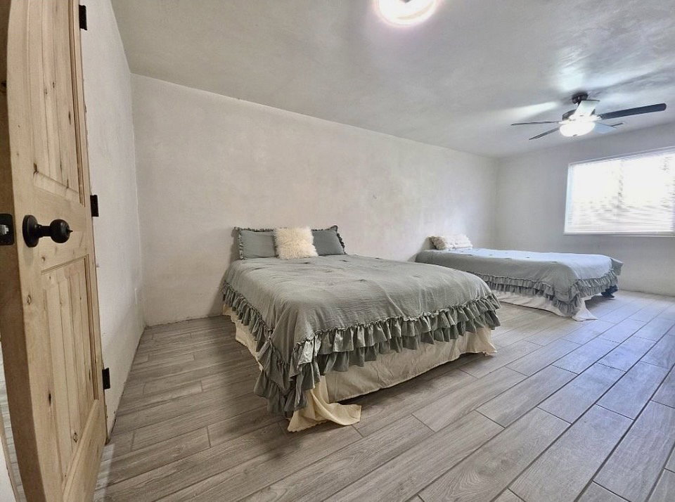 Spacious room featuring wood-finish flooring, a ceiling fan with integrated lighting, a flush-mount ceiling light, a window with horizontal blinds, and a rustic wood door with black hardware