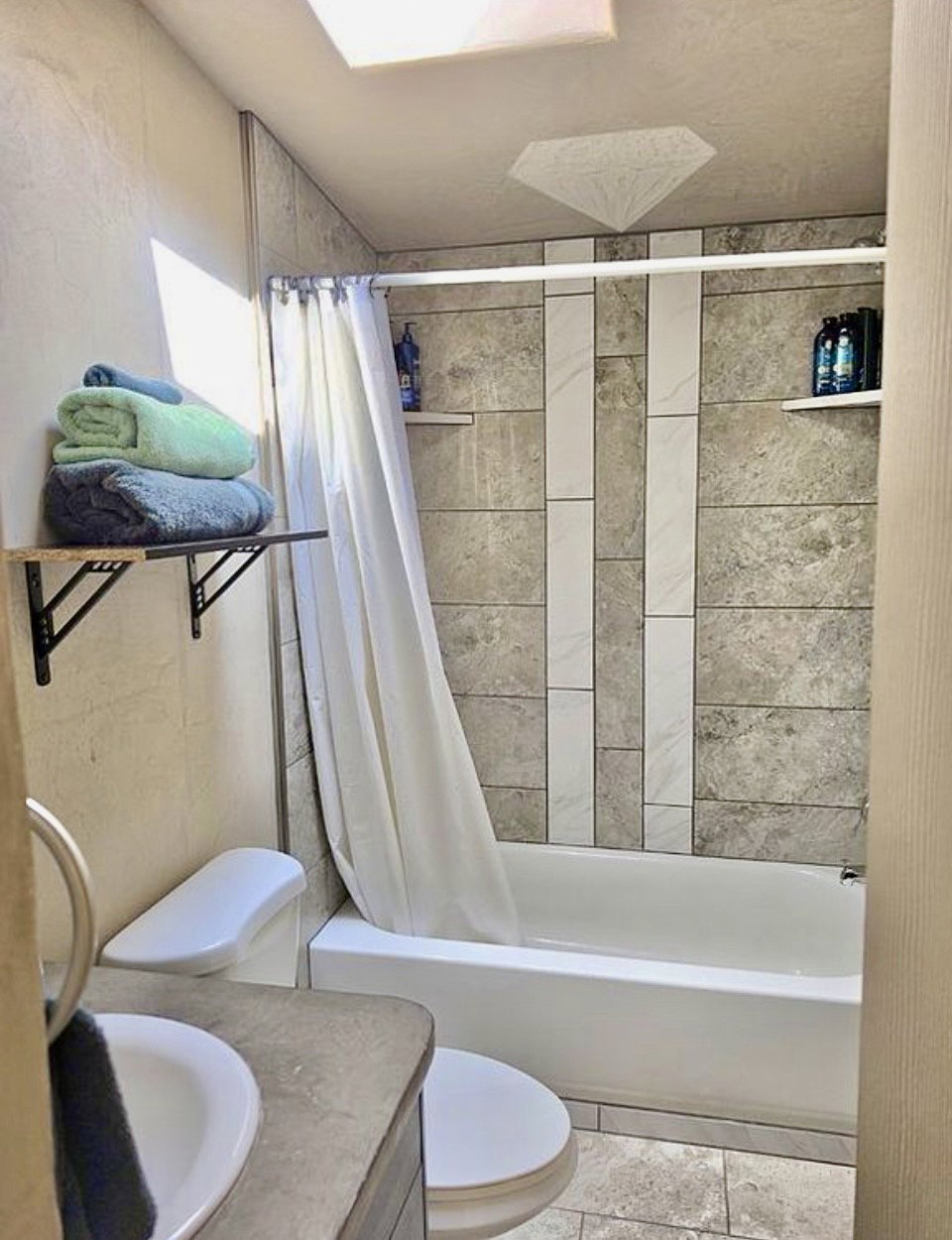 Bathroom featuring a white bathtub, tiled shower surround with accent strips, built-in shelving, a skylight, and a vanity with an integrated sink