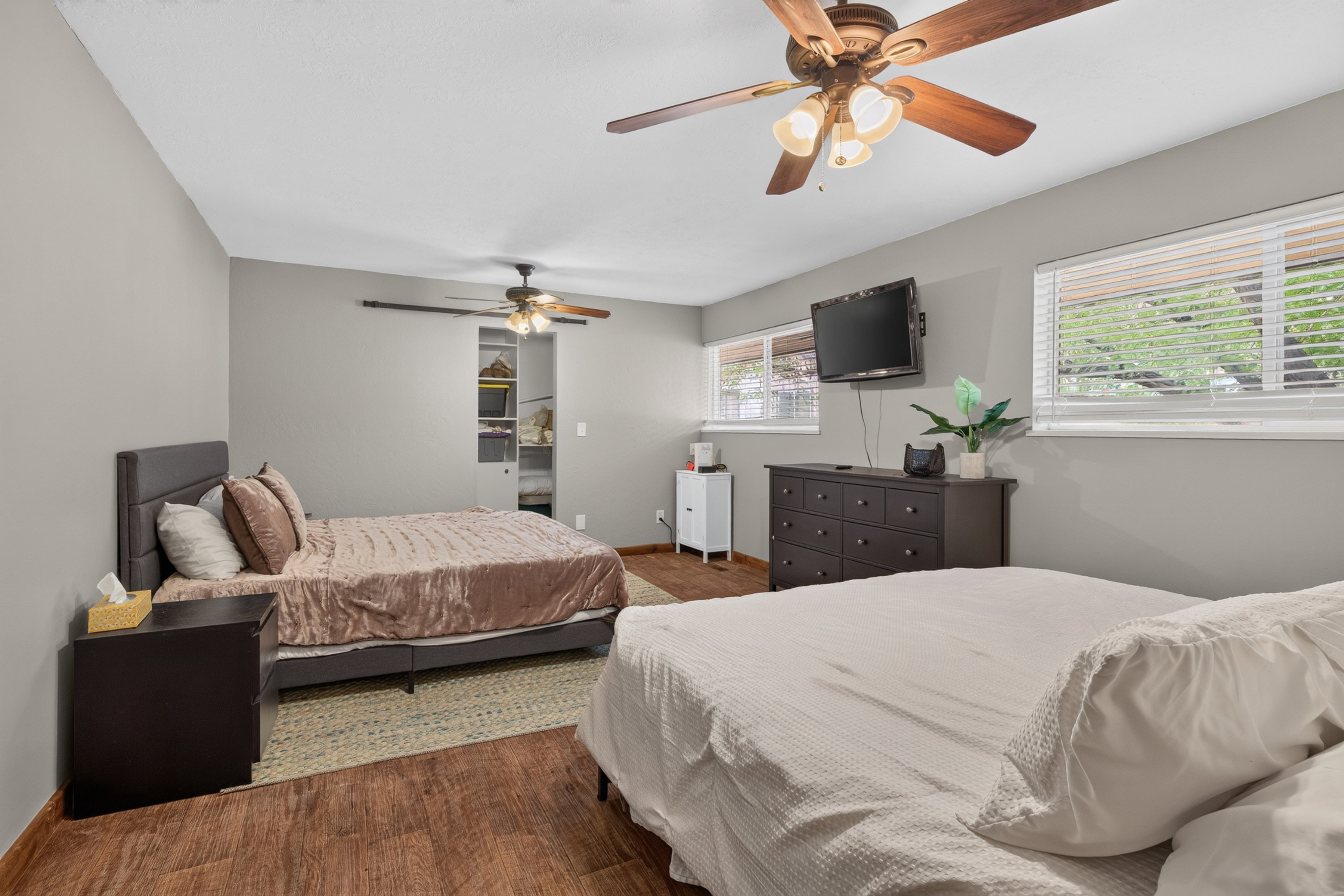 Spacious bedroom featuring wood-finish flooring, two large windows with blinds, and two ceiling fans with integrated lighting
