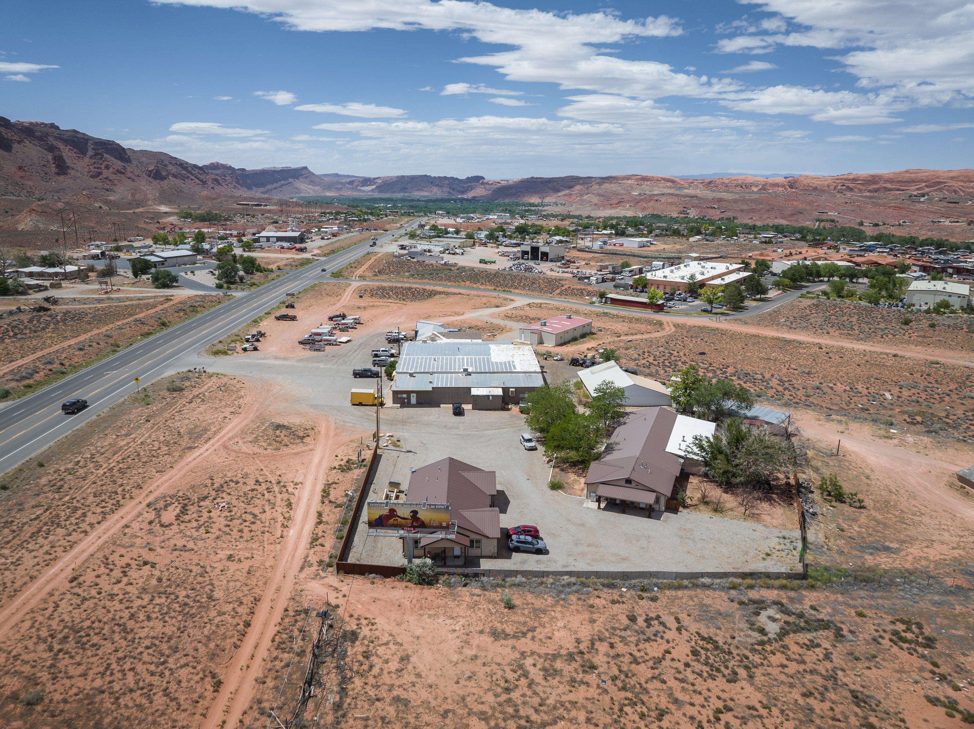 Commercial property featuring multiple buildings with brown roofs, a large gravel parking area, and a fenced perimeter