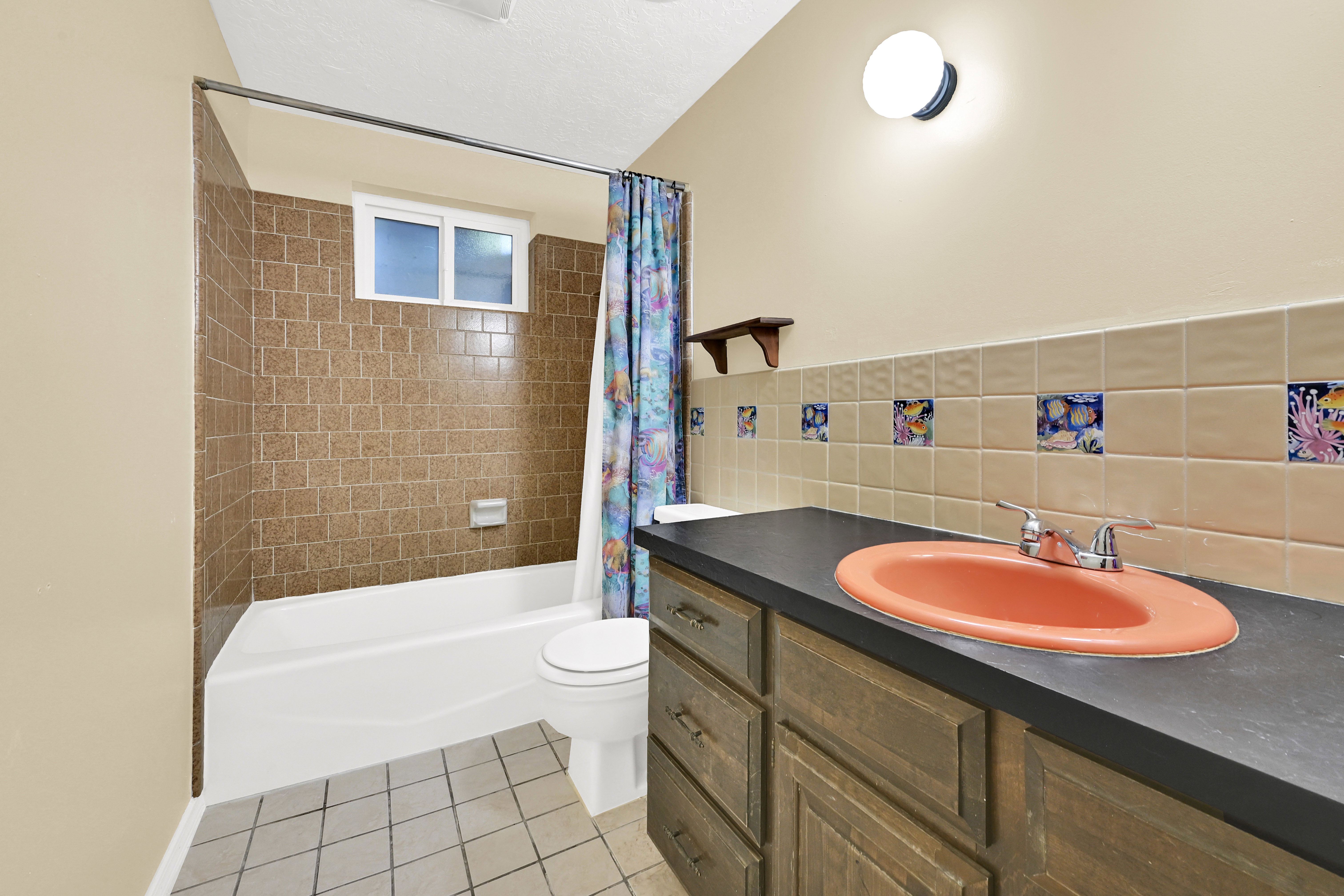 Bathroom featuring a white bathtub, brown tiled shower surround, window, single vanity with a dark countertop and orange basin, and neutral floor tiles