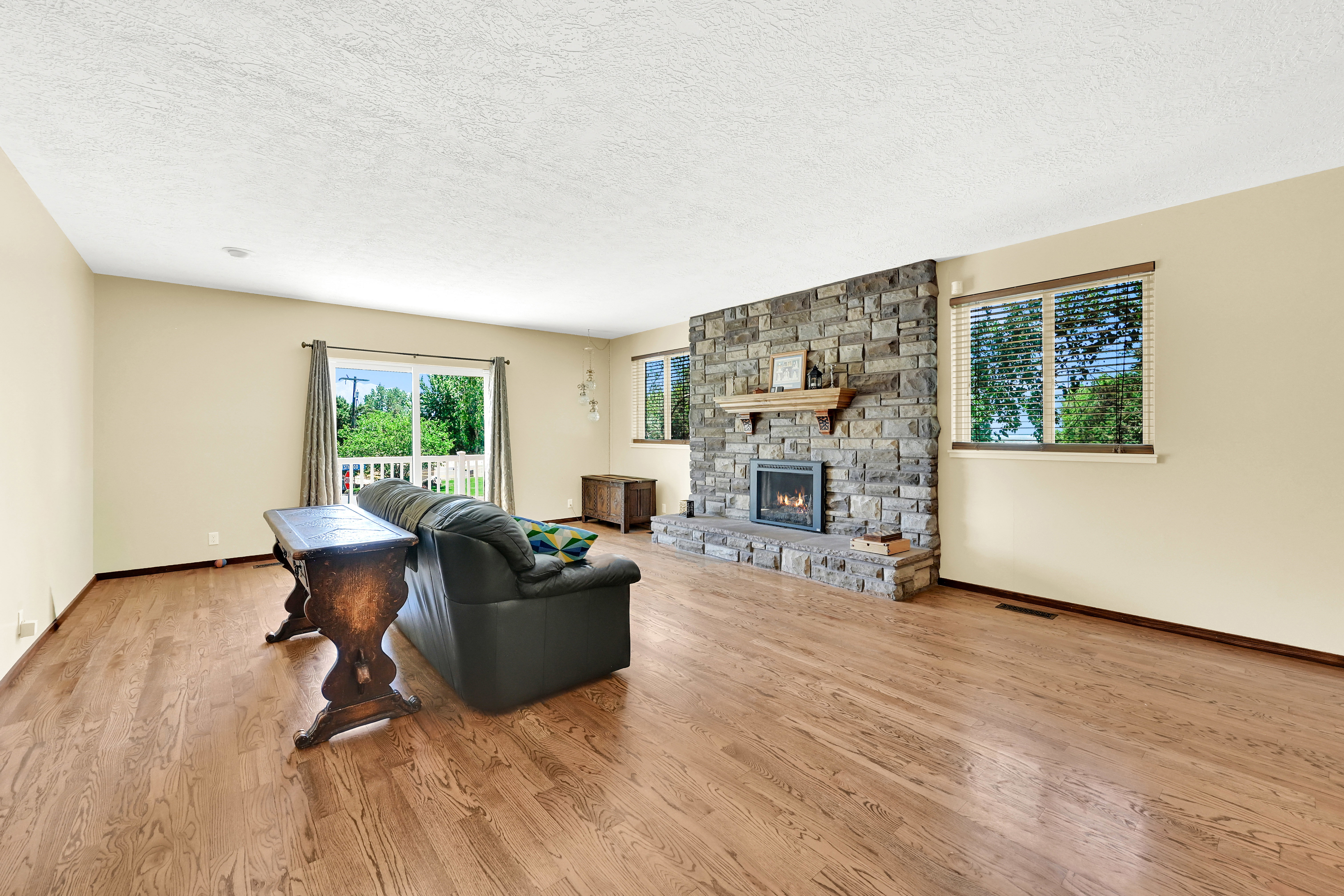 Spacious living area featuring wood-finish flooring, a stone-clad fireplace with a wood mantel, and multiple windows with horizontal blinds