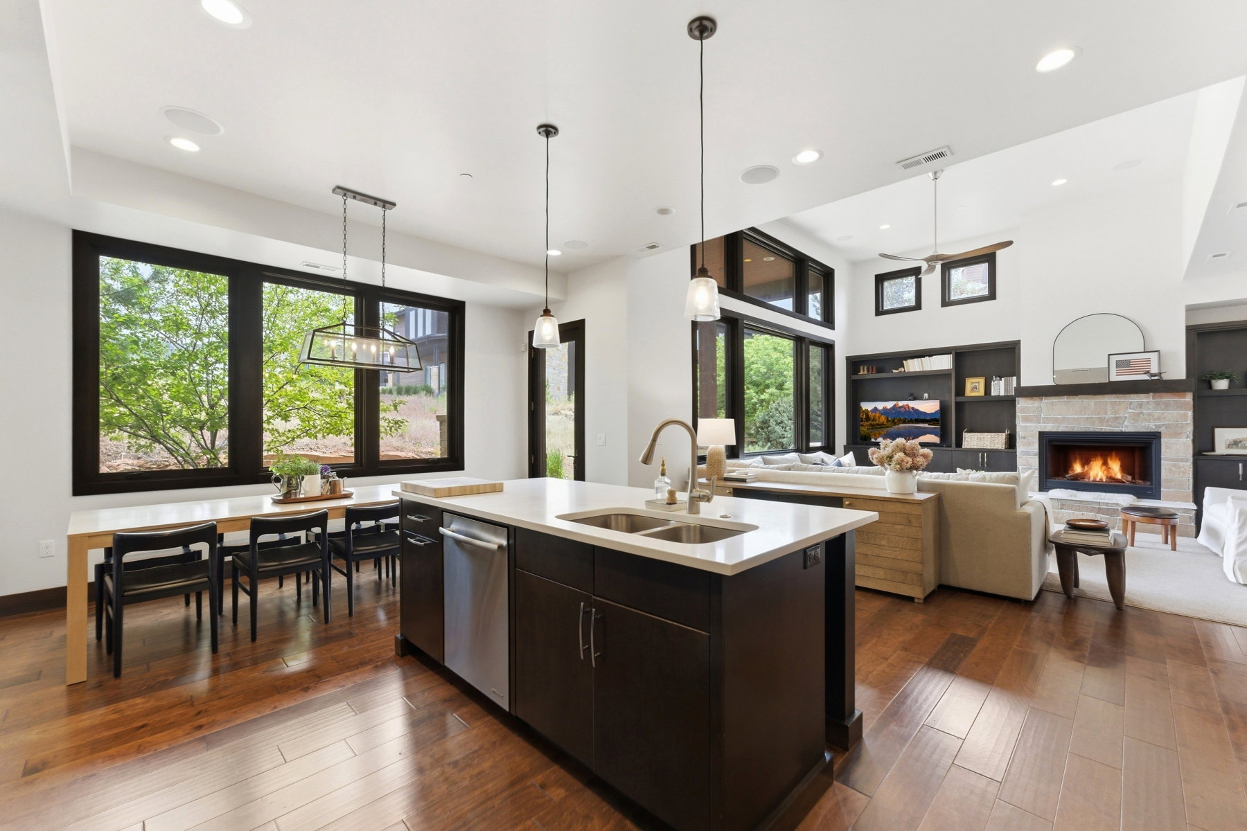 Open-concept living space featuring wood-finish flooring, a kitchen island with dual sinks, a built-in fireplace with stone surround, and an integrated entertainment unit with shelving