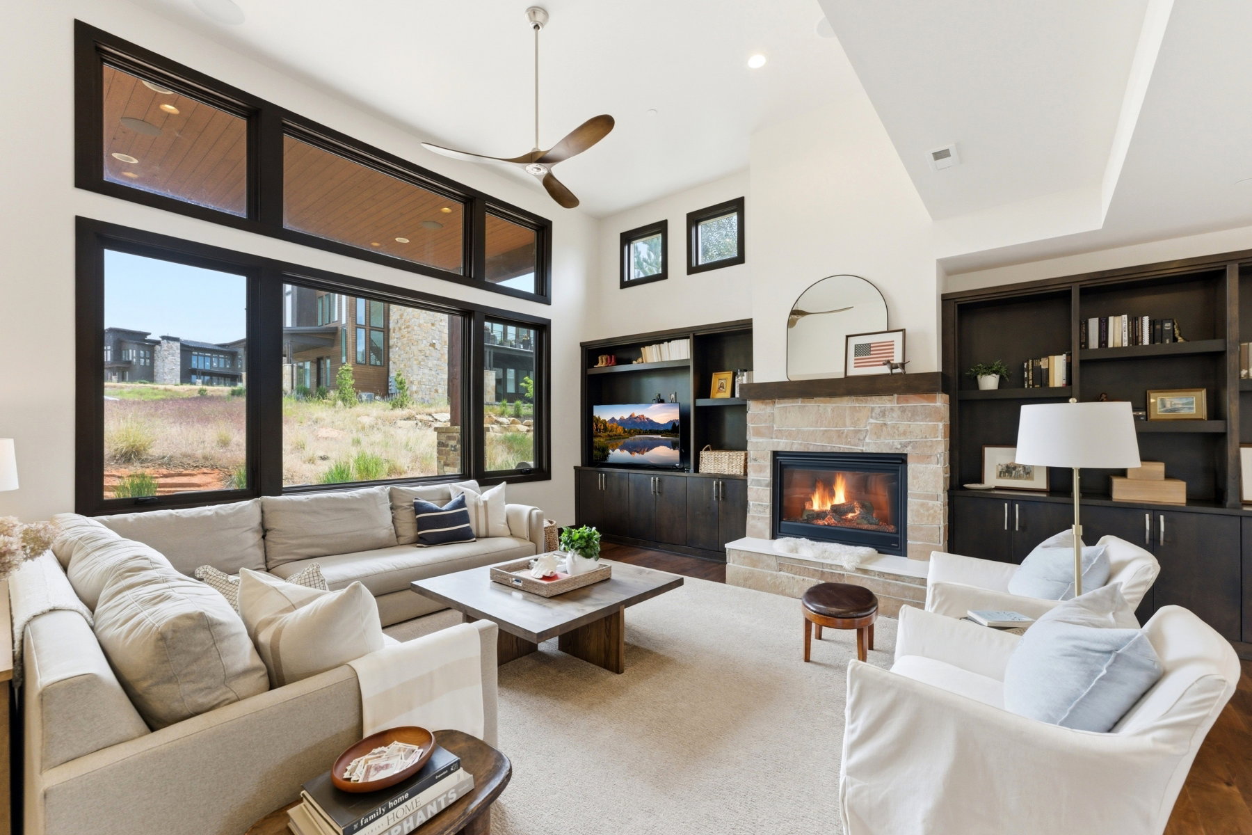 Spacious living area featuring a stone-clad fireplace, integrated dark wood-finish cabinetry and shelving, high ceilings, large dark-framed windows, and a ceiling fan