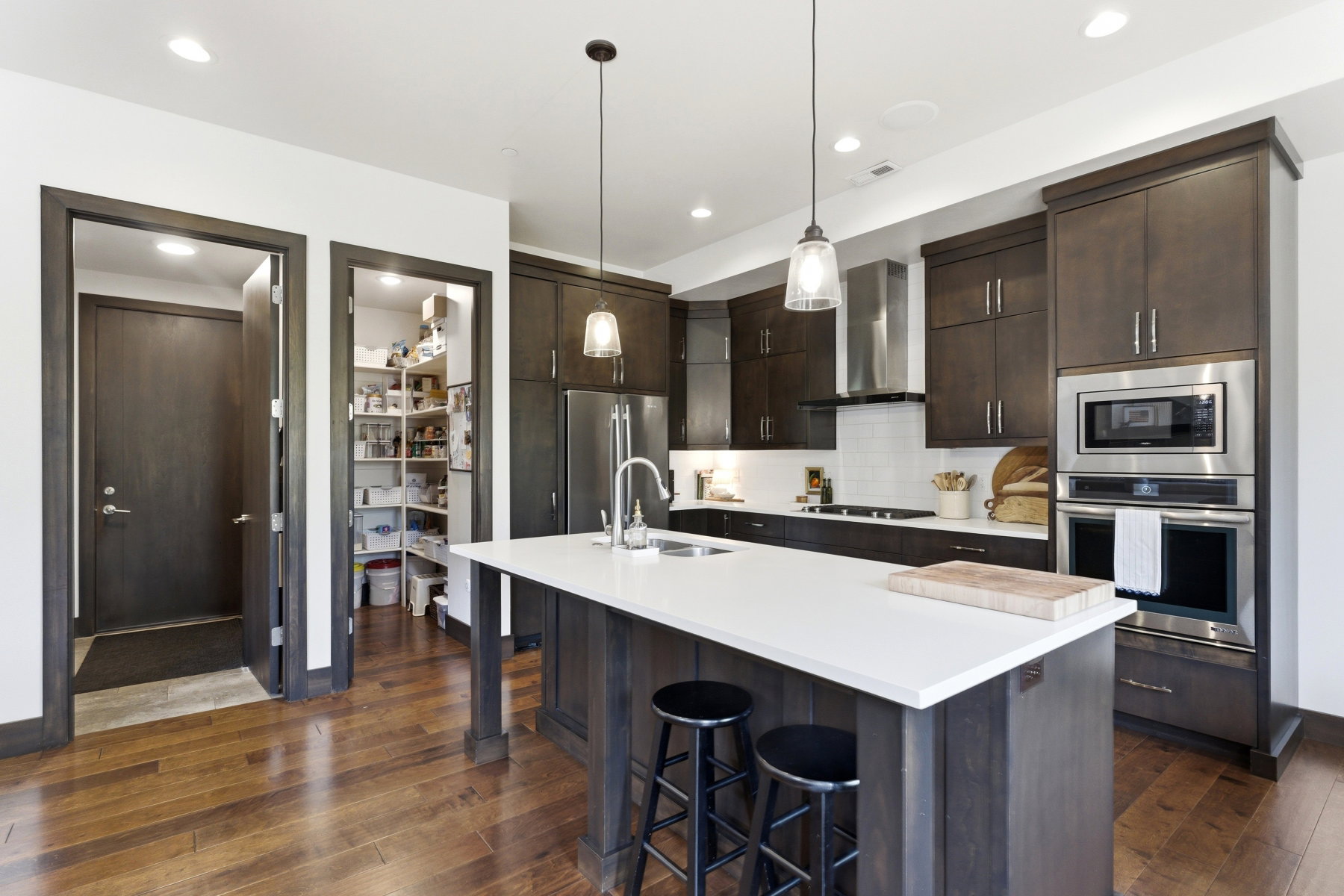 Contemporary kitchen featuring an island with a white countertop, dark wood-finish cabinetry, stainless steel appliances, a tile backsplash, and wood-finish flooring