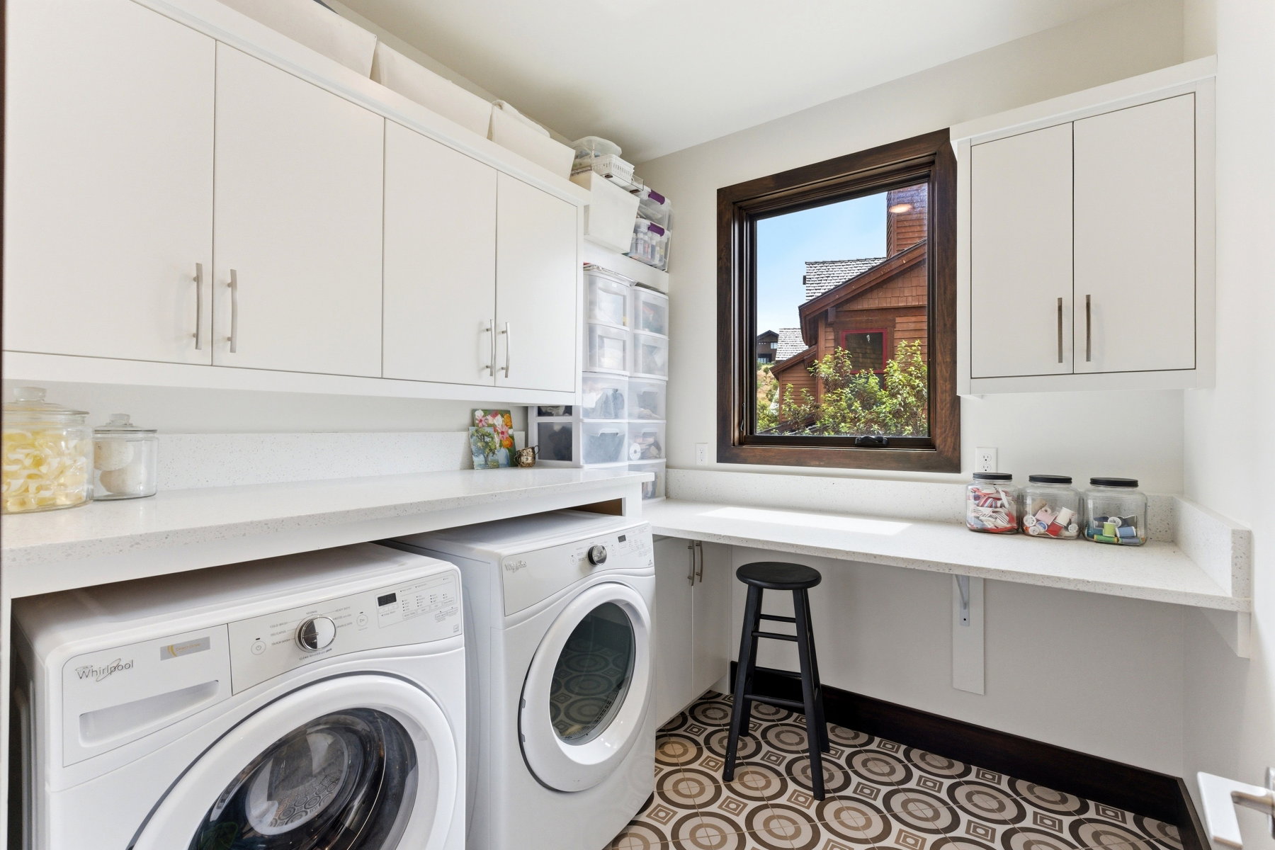 Dedicated utility space featuring white cabinetry with brushed nickel hardware, solid surface countertops, a dark wood-framed window, and patterned tile flooring