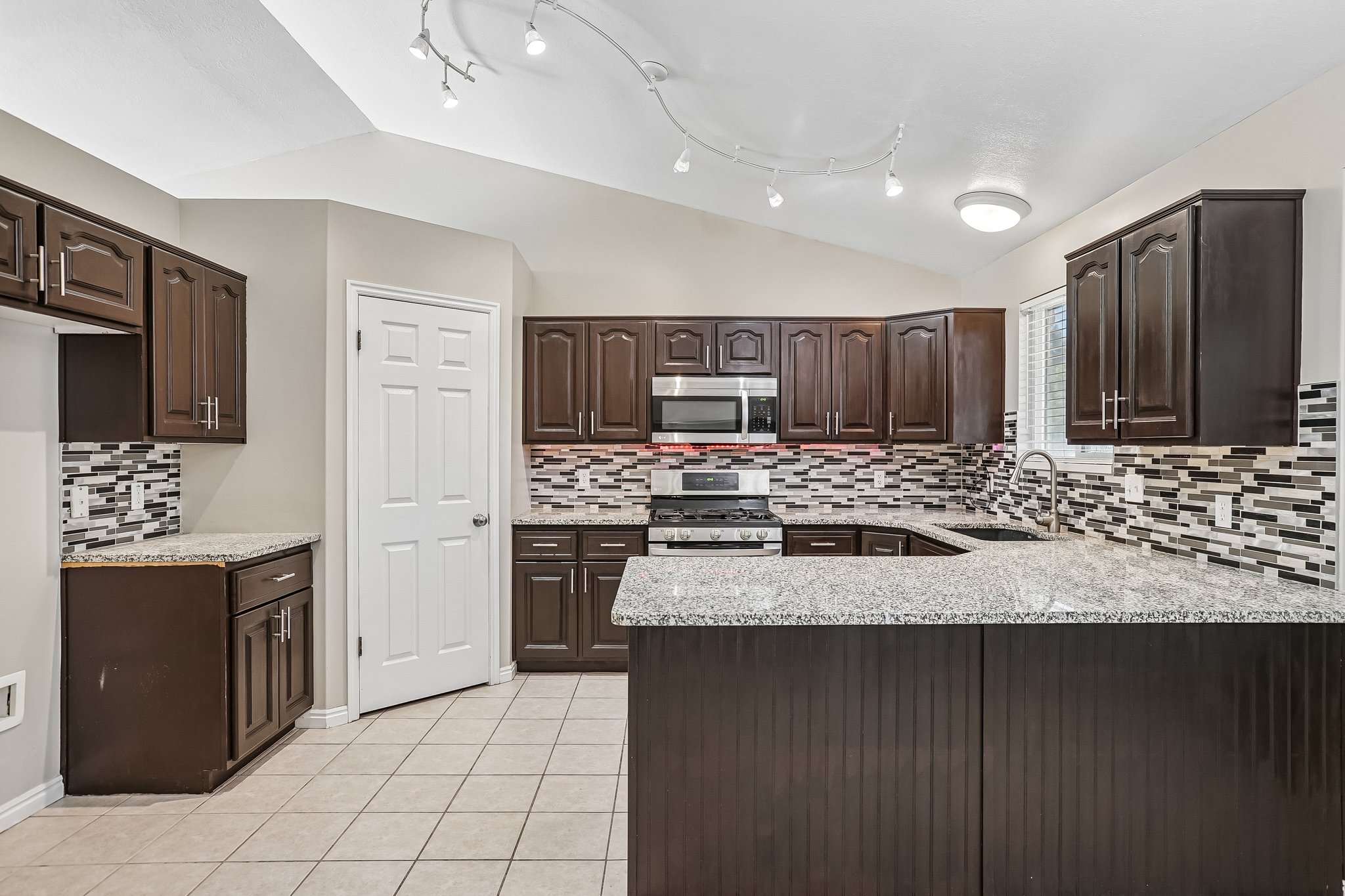 Kitchen featuring dark wood cabinetry, light-toned countertops, a mosaic tile backsplash, stainless steel appliances, and vaulted ceilings with track lighting