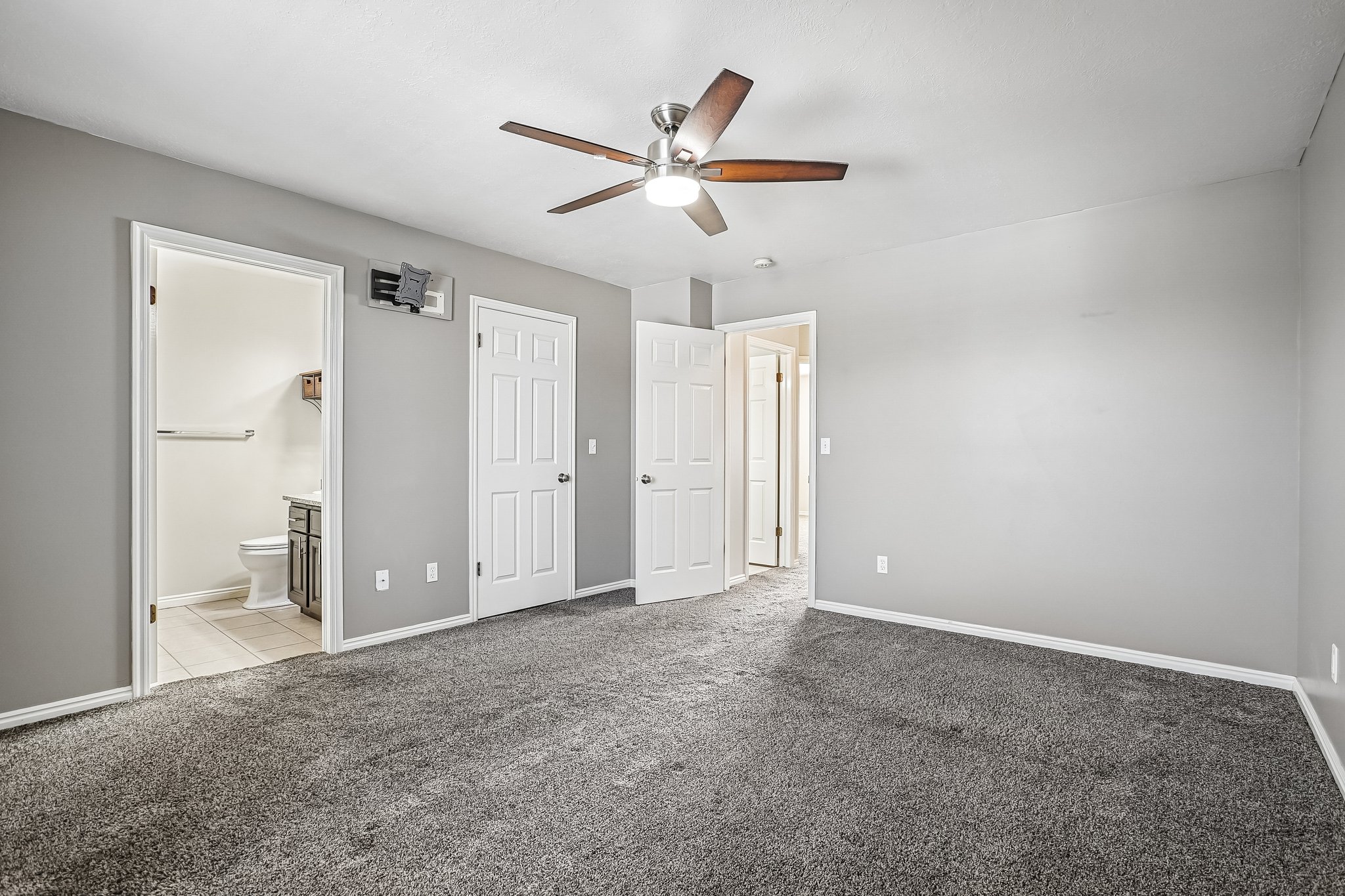 Carpeted room with light gray walls, a ceiling fan with wood-finish blades, and multiple white paneled doors