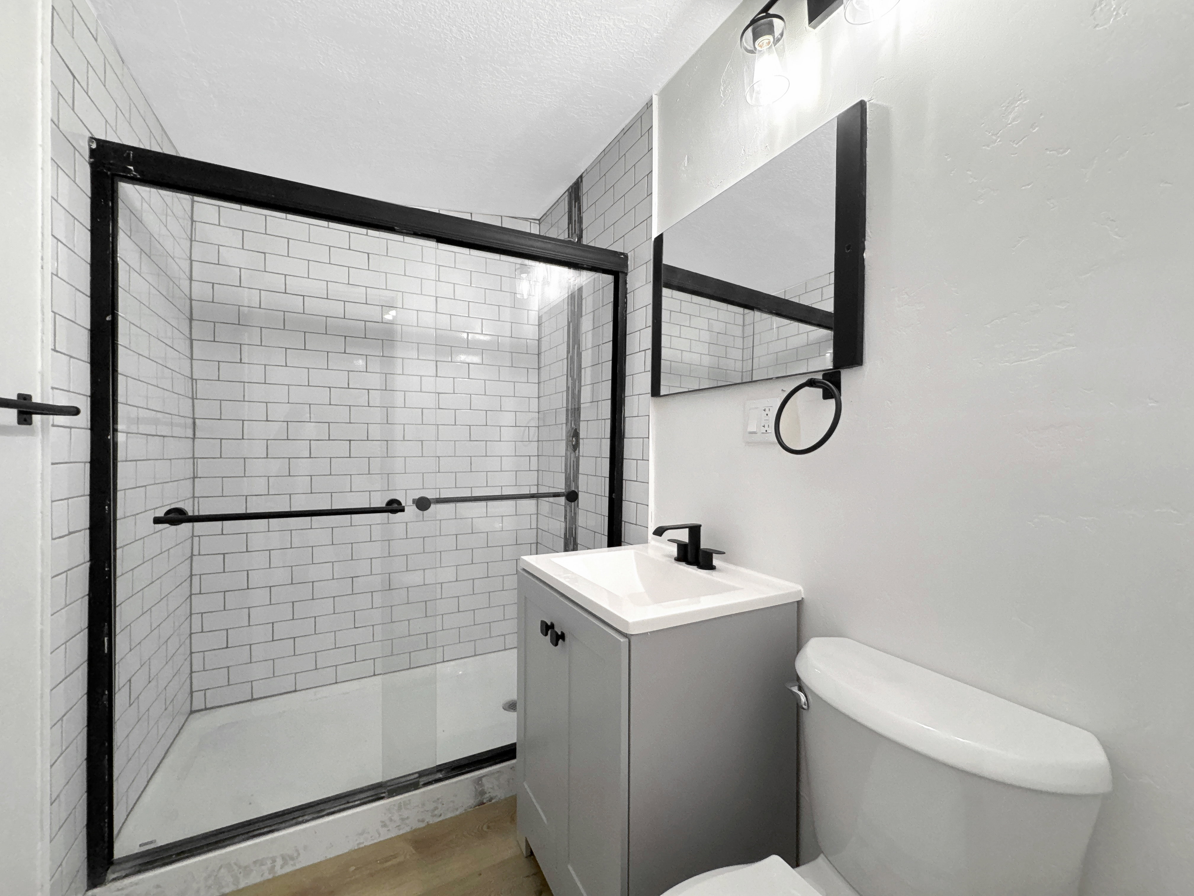 Bathroom featuring a shower with white subway tile surround and black-framed glass enclosure, a vanity with integrated sink and black faucet, and wood-finish flooring.  in detached ADU