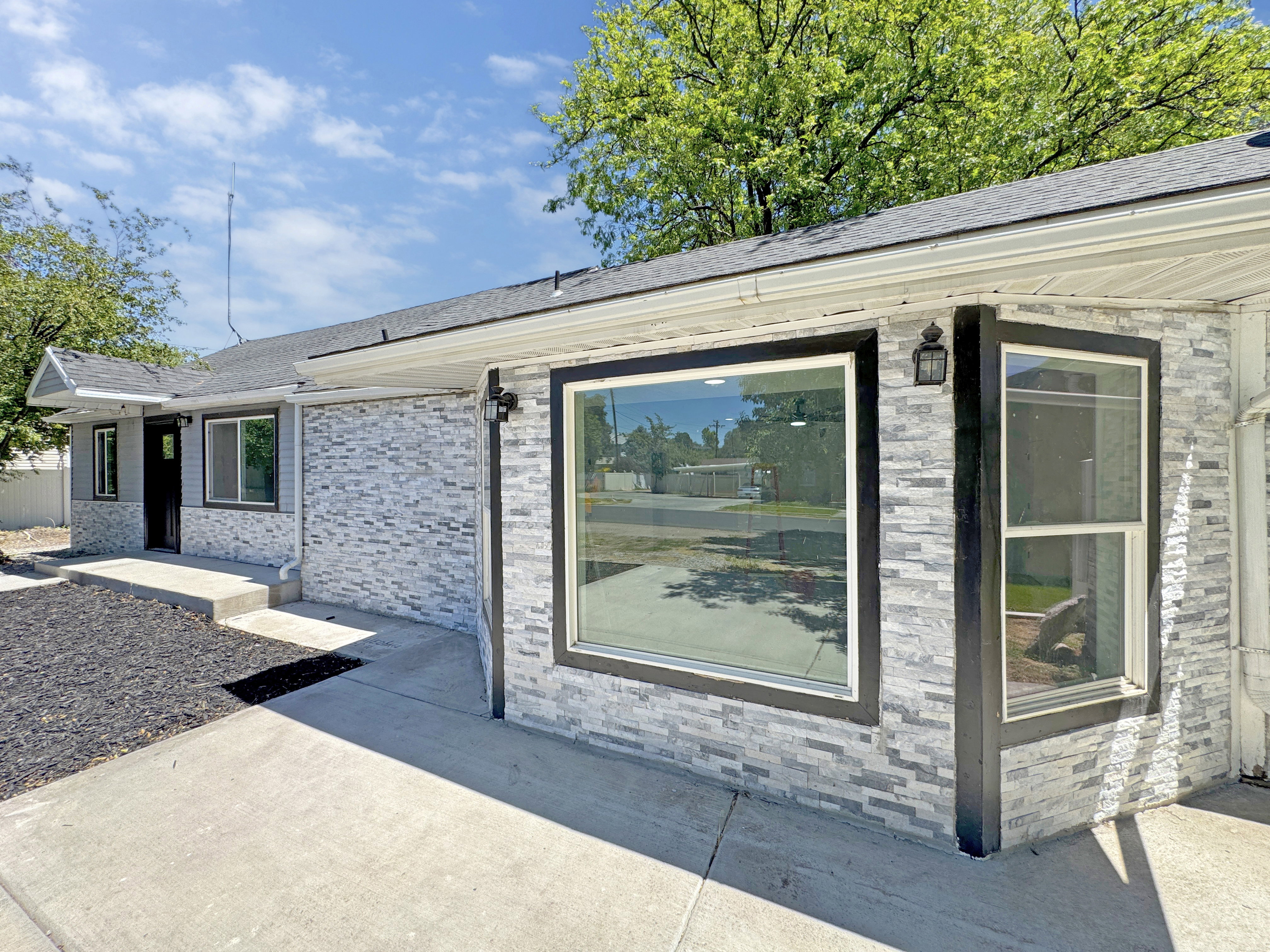Exterior featuring grey stacked stone veneer, a dark shingle roof, dark window trim, and exterior sconce lighting