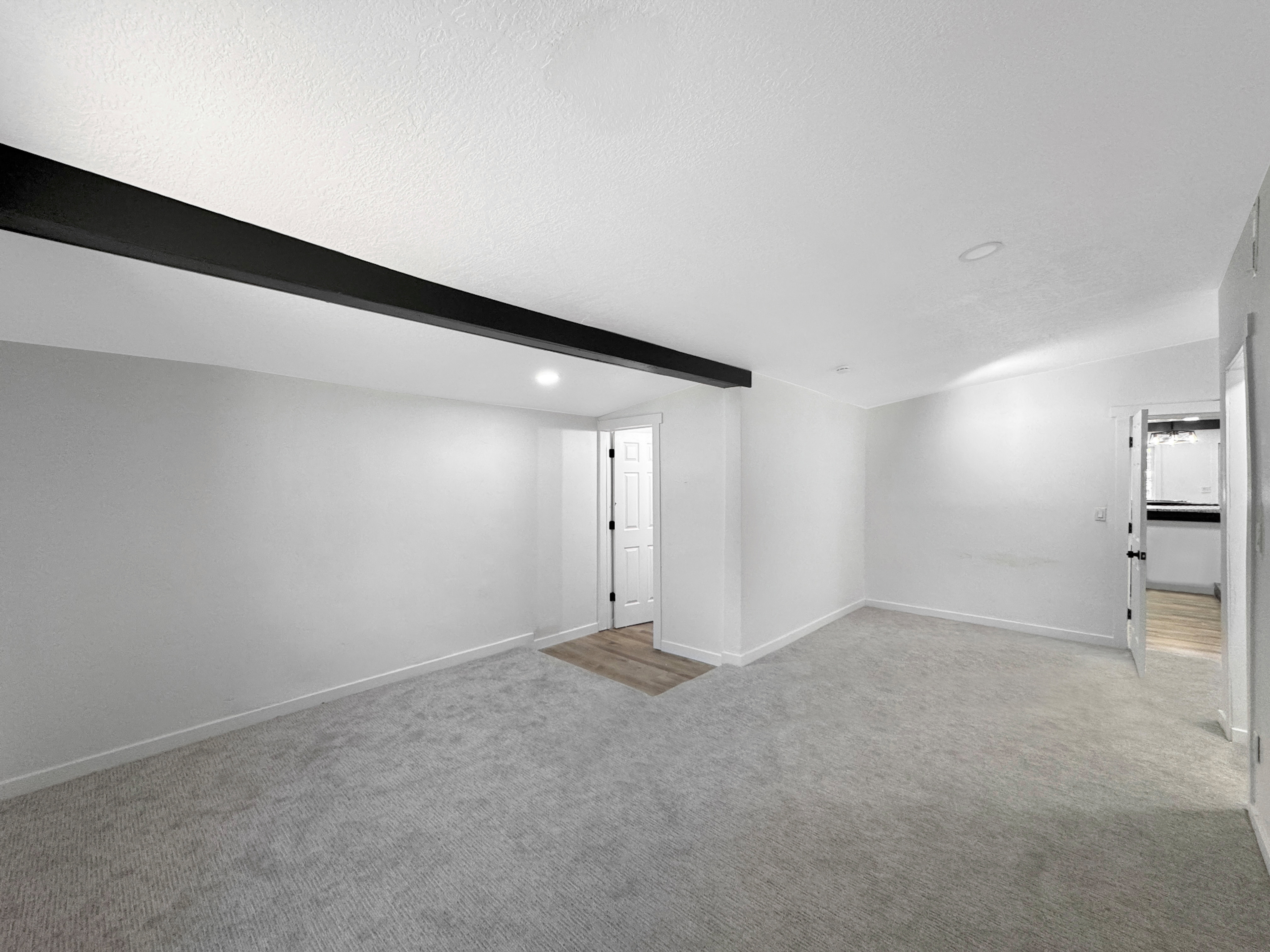 Spacious room featuring light gray carpeting, crisp white walls, and a prominent black ceiling beam